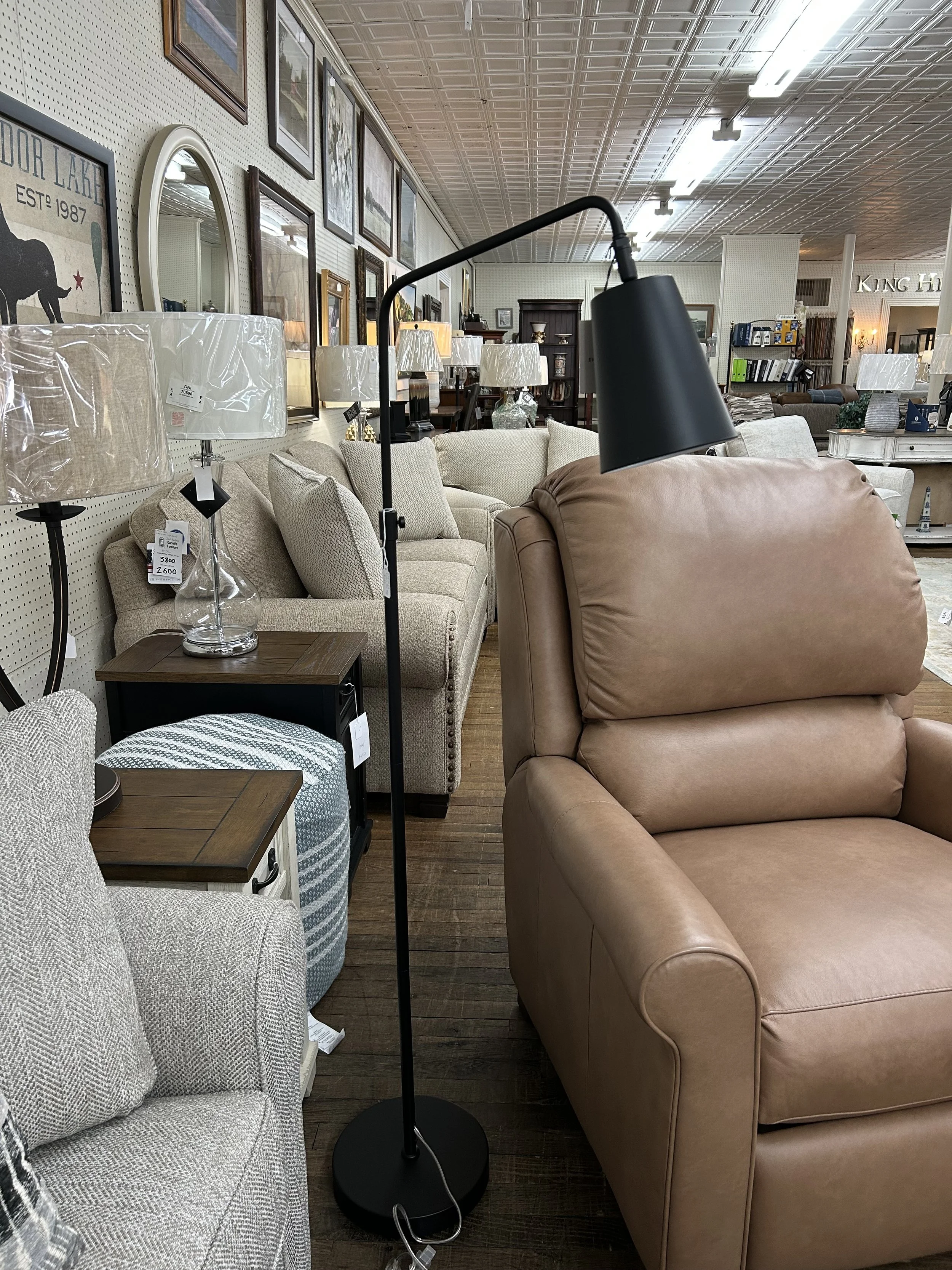 A black standing floor lamp with a curved arm and black lampshade standing among beige and light-colored sofas and furniture in a furniture store.