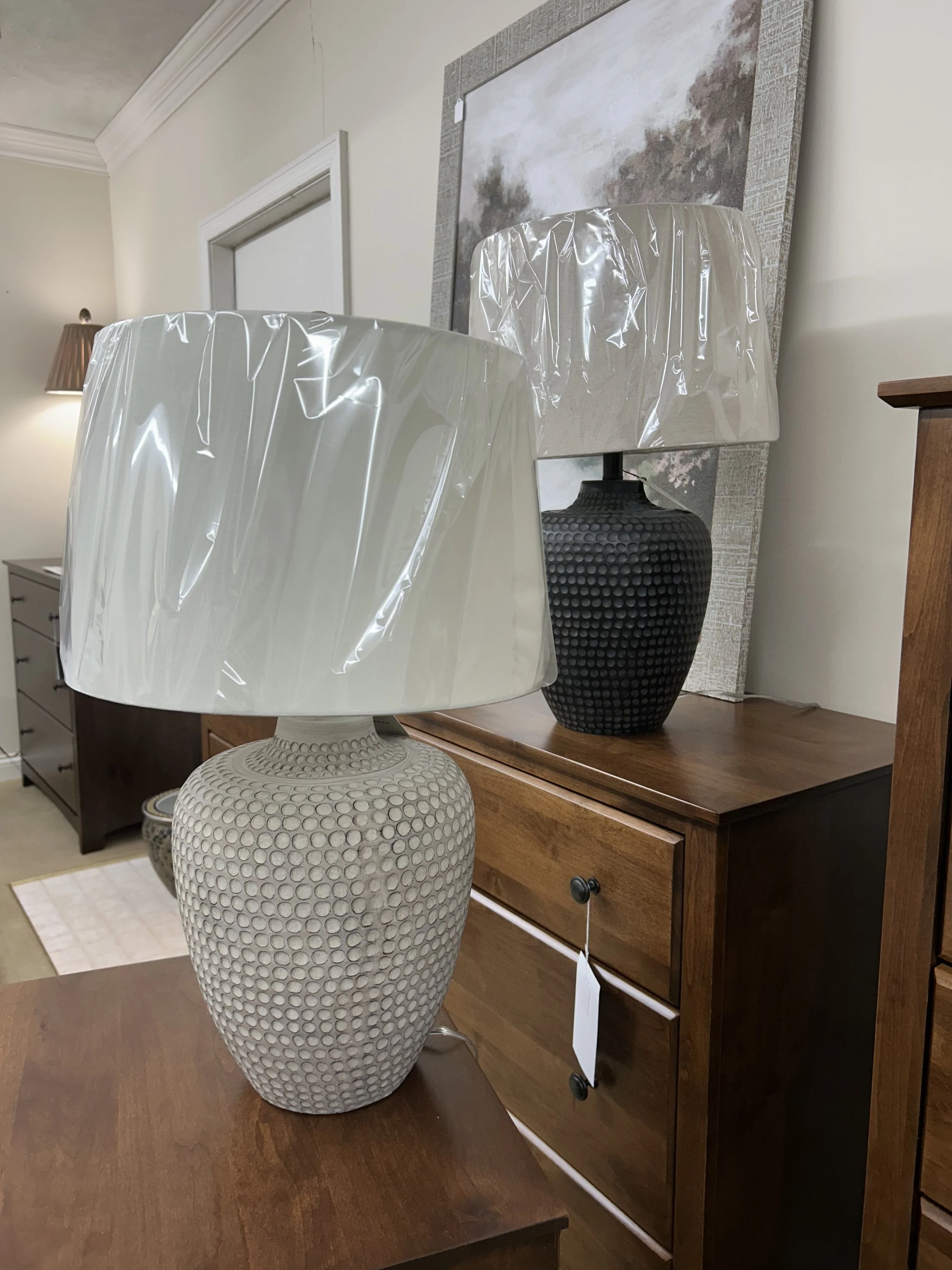 Two decorative table lamps with large white and black textured bases wrapped in plastic, on wooden furniture in a room with wall art and a side table.