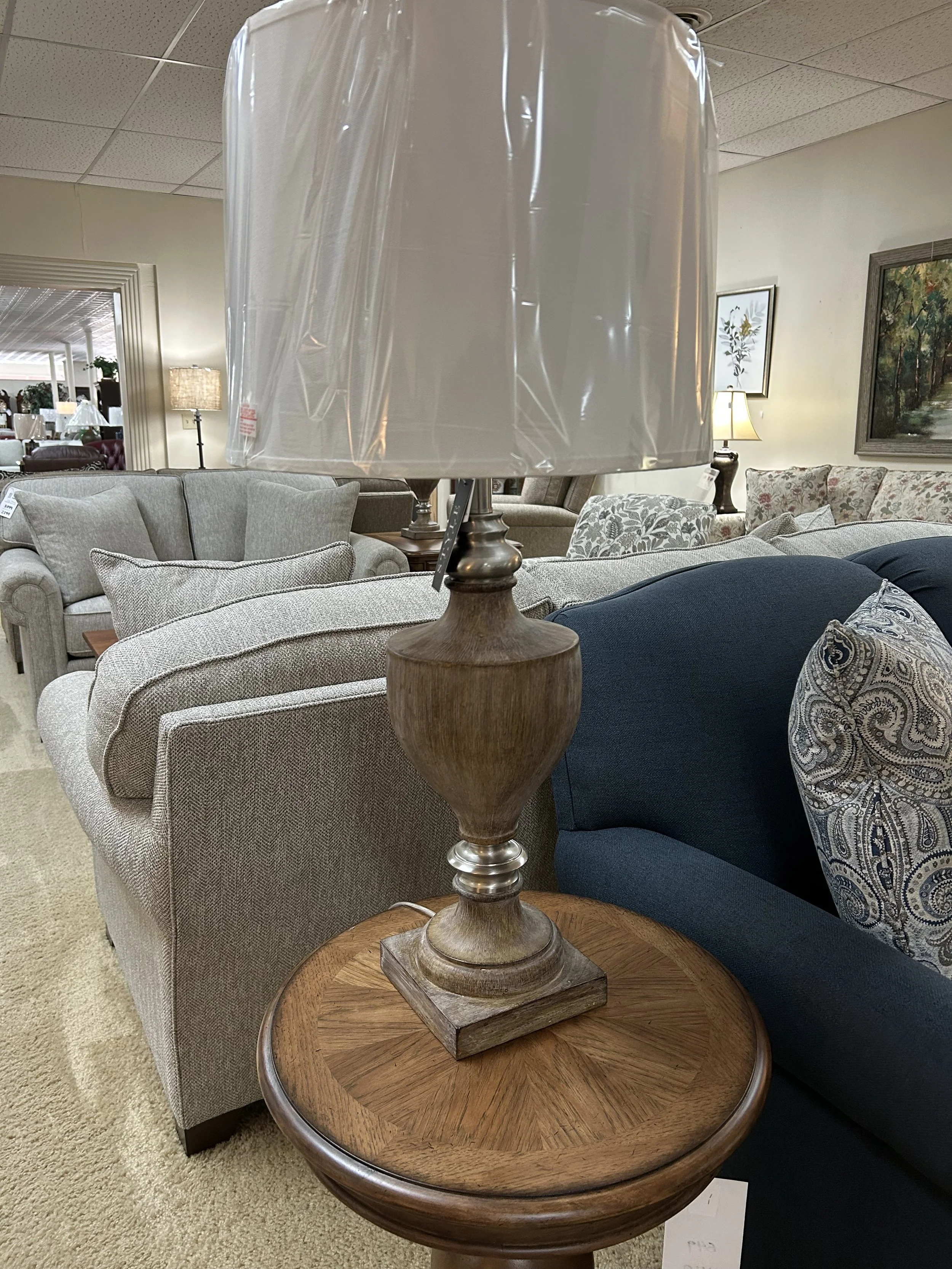 Wooden table lamp with a plastic protective cover on its lampshade, placed on a round wood table in a furniture store.