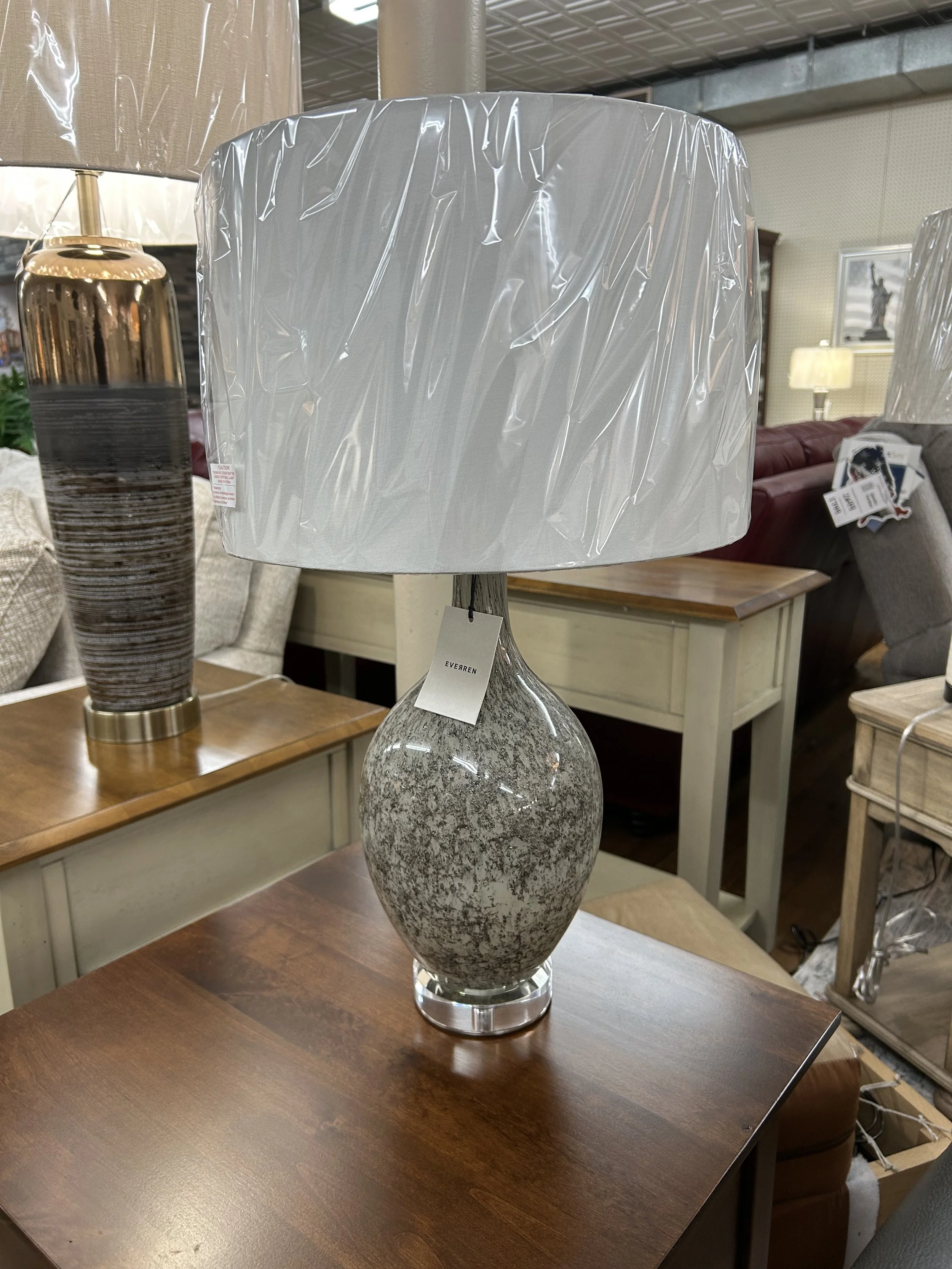 A decorative table lamp with a stone-textured ceramic base and a large cylindrical white lampshade covered in plastic wrapping, placed on a wooden table in a furniture store.