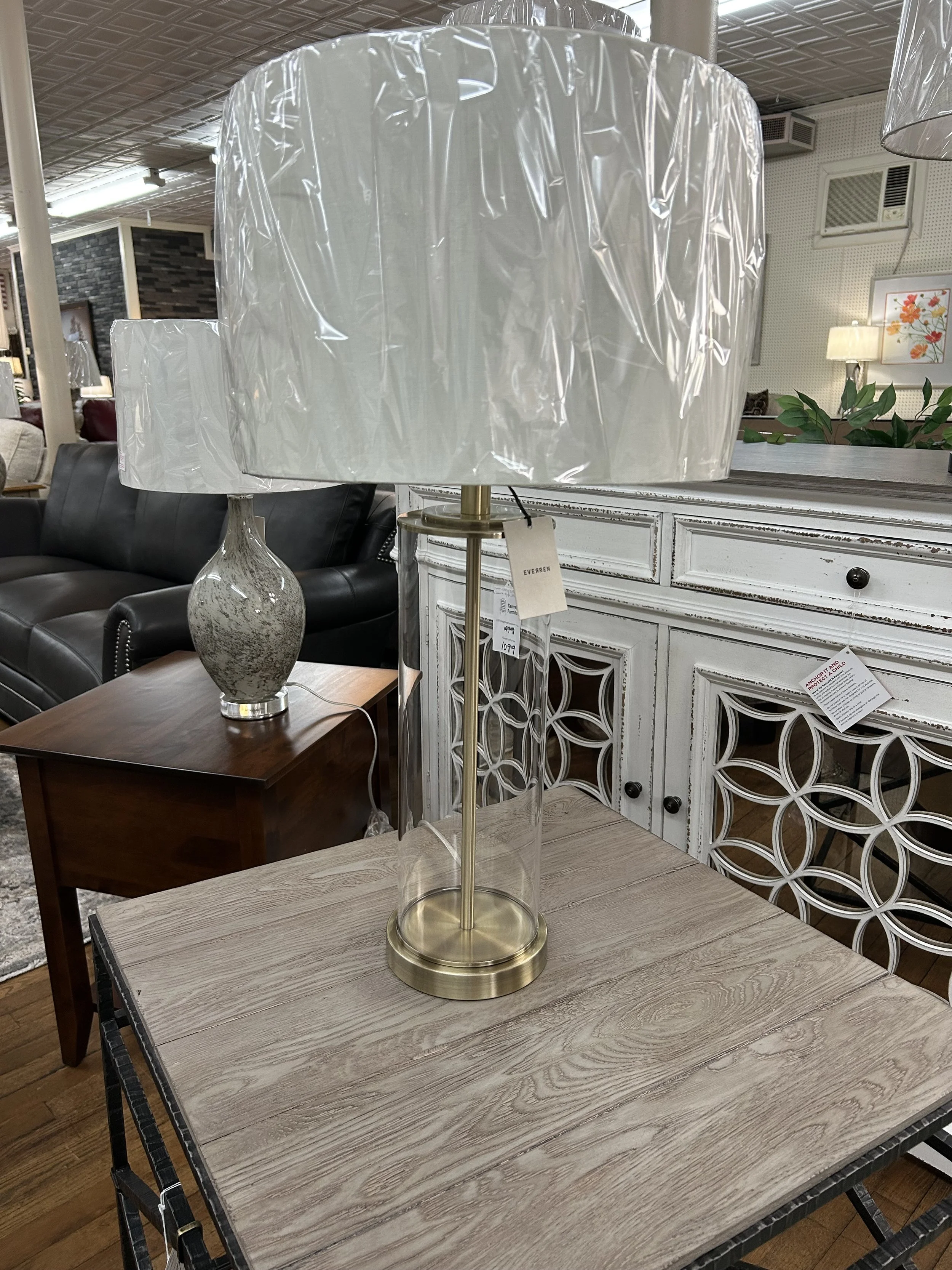 A table lamp with a large, off-white, plastic or fabric shade and a tall, cylindrical, transparent glass base with gold-colored metal accents, placed on a light wood table in a furniture store.