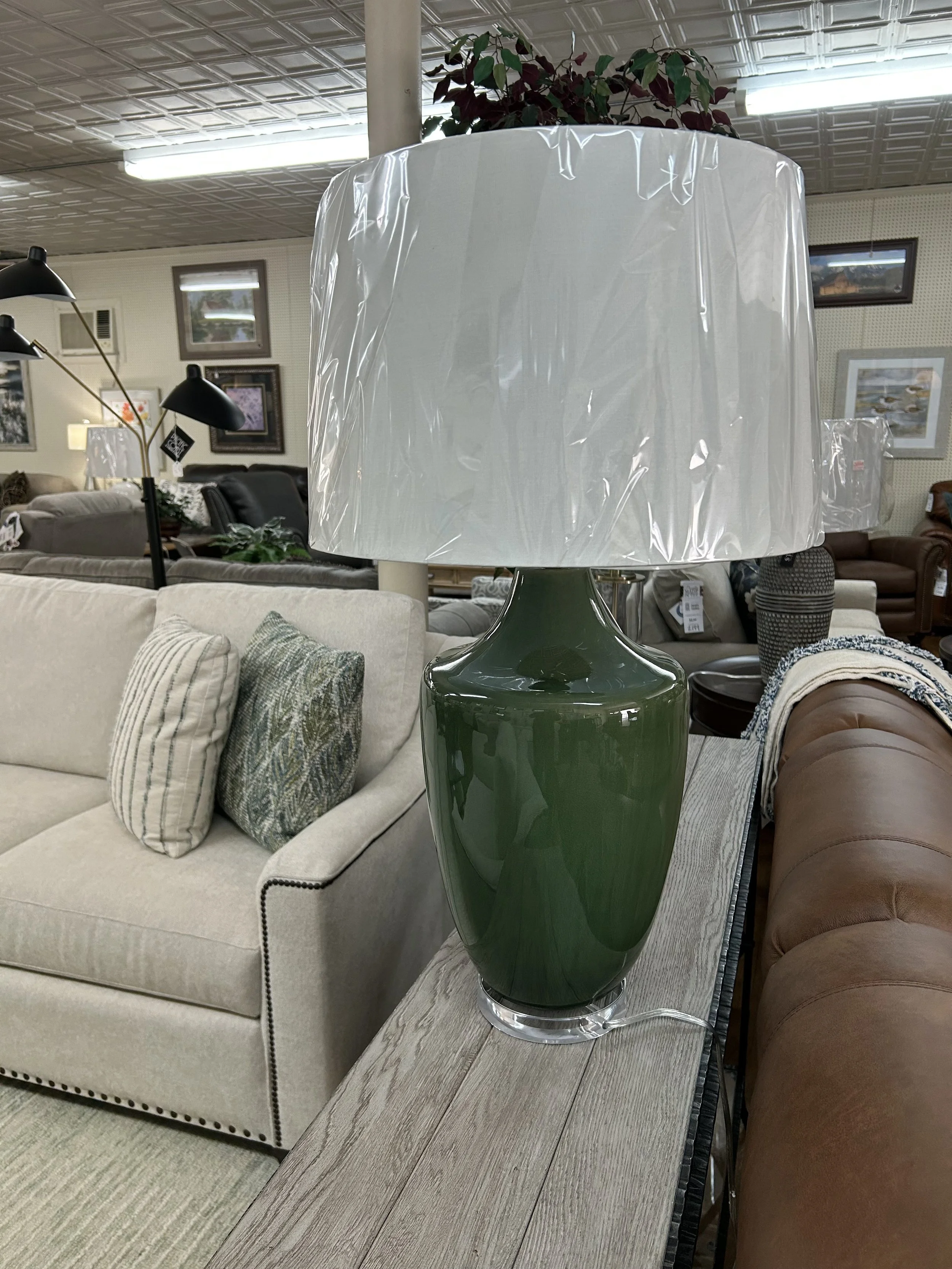 A green ceramic table lamp with a white lampshade wrapped in plastic, placed on a wooden surface in a furniture store.