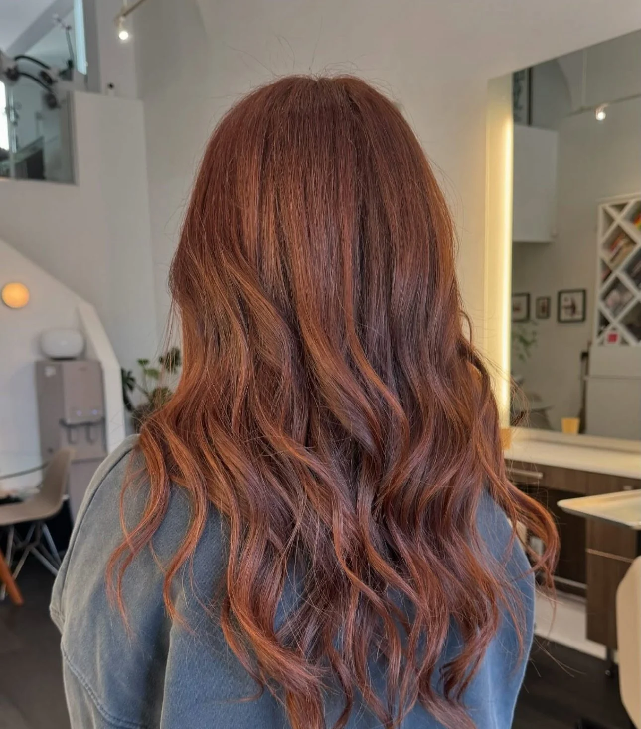 A woman with long, wavy, reddish-brown hair standing in a modern interior space.