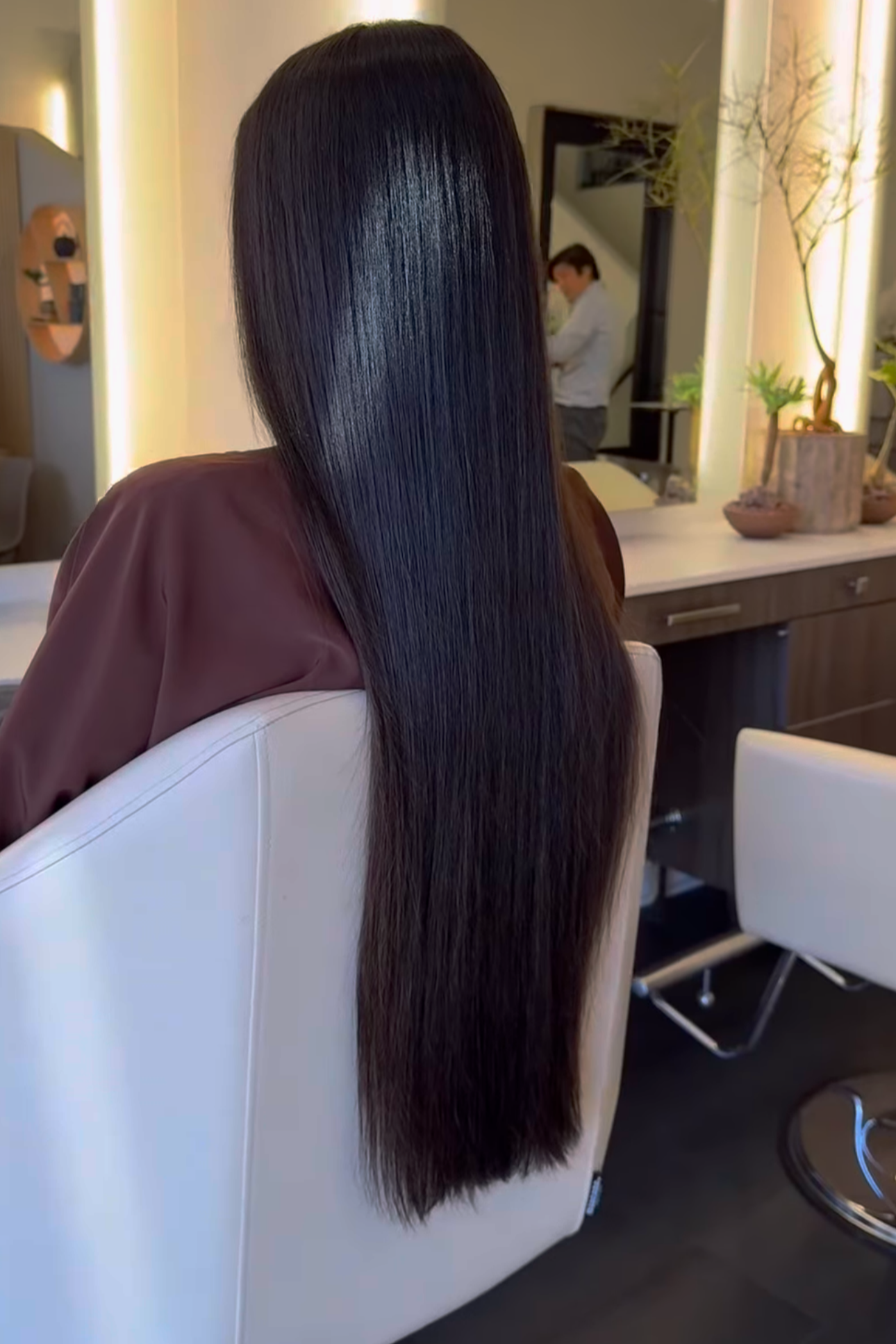 Close-up of a woman with long, straight, and shiny dark hair sitting in a salon chair, facing away from the camera.