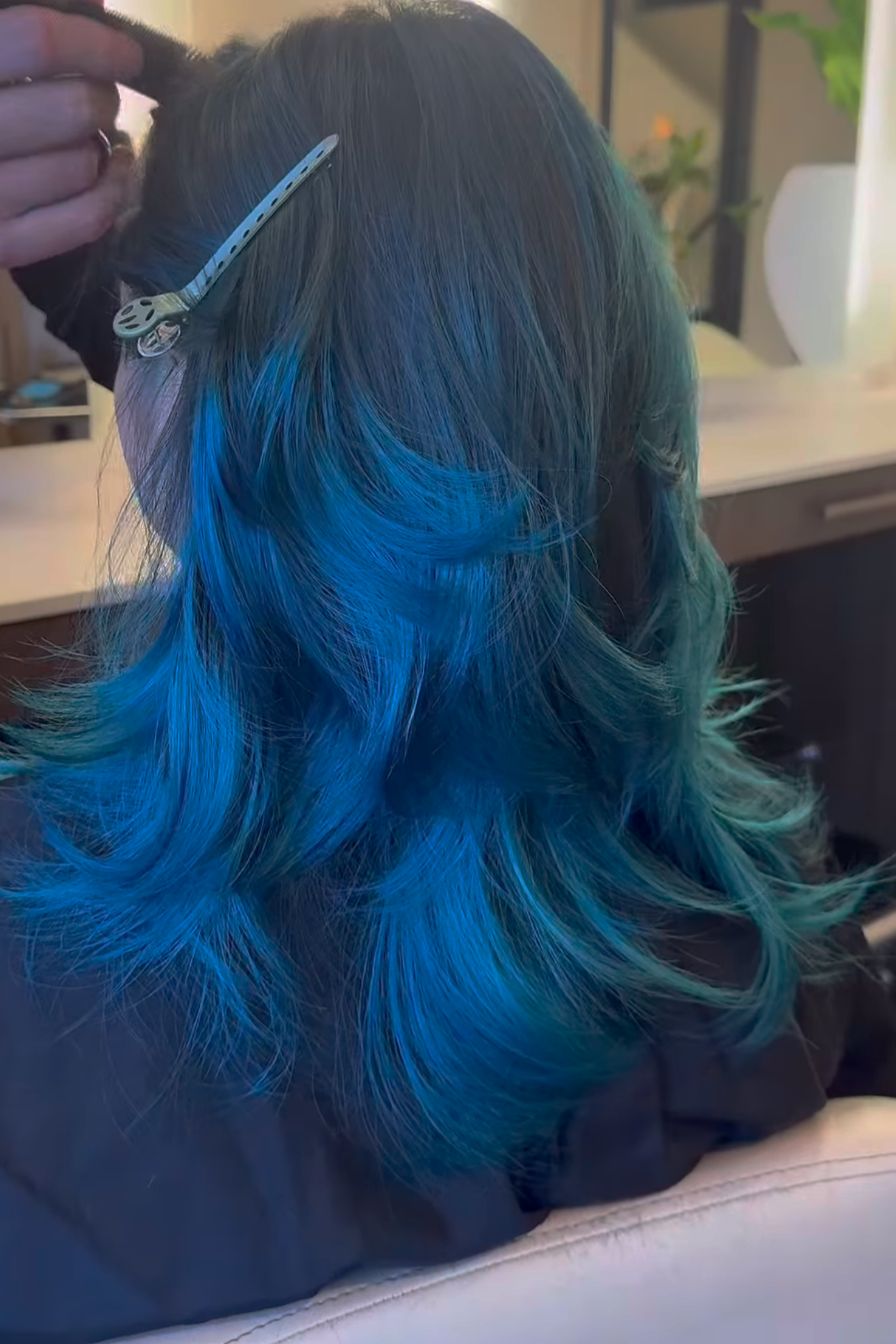 Woman getting her hair styled with blue coloring, seated in a salon chair, with hairstylist working on her hair.