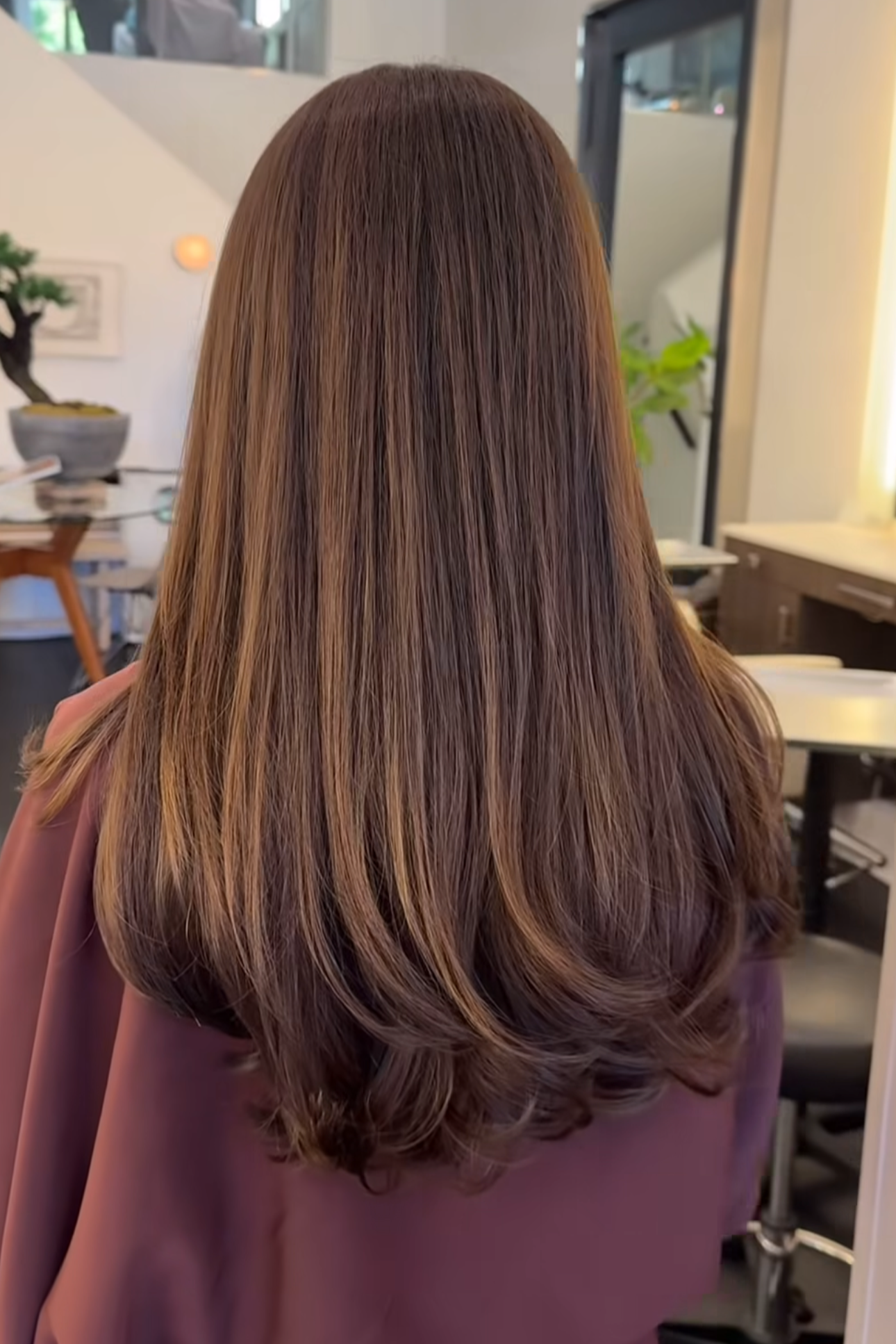 Back of a woman with long, smooth, brown hair styled with soft curls at the ends, sitting in a salon.