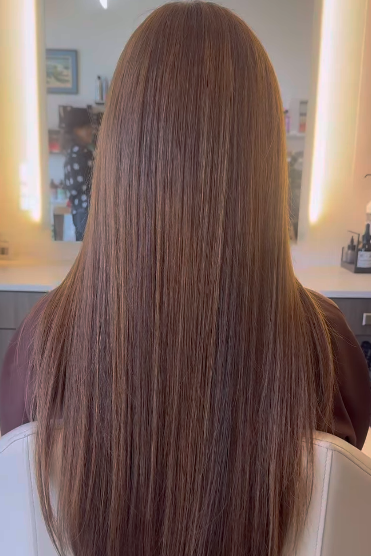 Back of a woman with long, straight, shiny red hair in a salon mirror reflection.