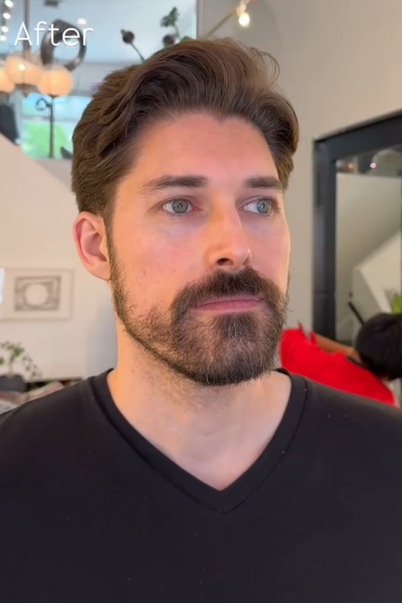A man with brown hair styled back, a full beard, and blue eyes looking slightly to the right. He is wearing a black V-neck shirt. The background shows a hanging light fixture, some plants, and a person in a red shirt facing away.