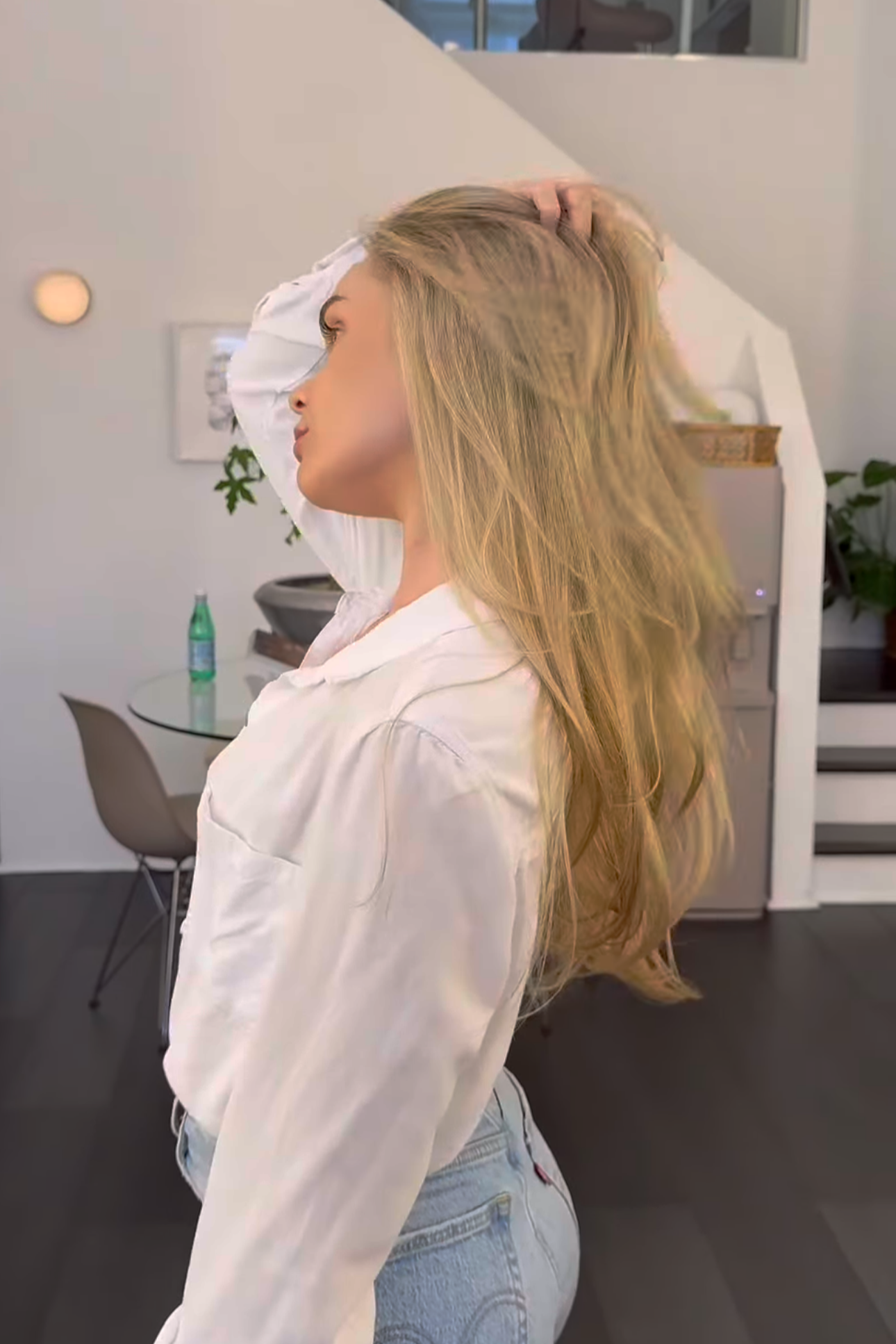 A woman with long blonde hair wearing a white shirt and light-wash jeans is posing indoors, with her left hand running through her hair and her eyes closed, in a modern room with minimalist décor.