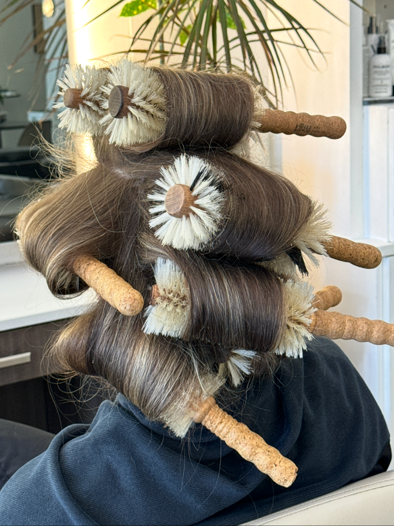 A person with brown hair getting their hair styled with large curlers and brushes in a salon.