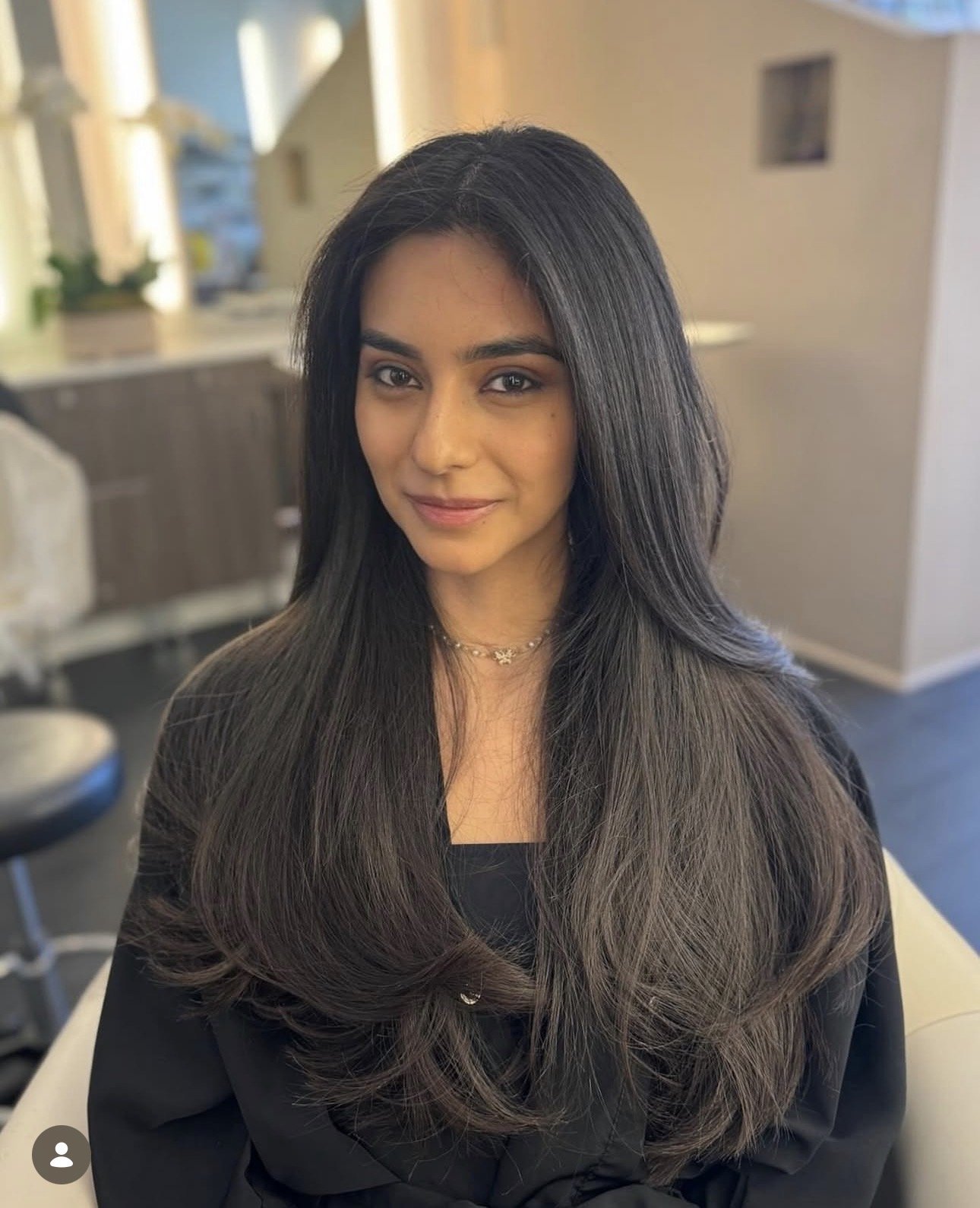 A young woman with long dark hair, wearing a black top and a dainty necklace, smiling at the camera in a room with neutral walls and some furniture in the background.