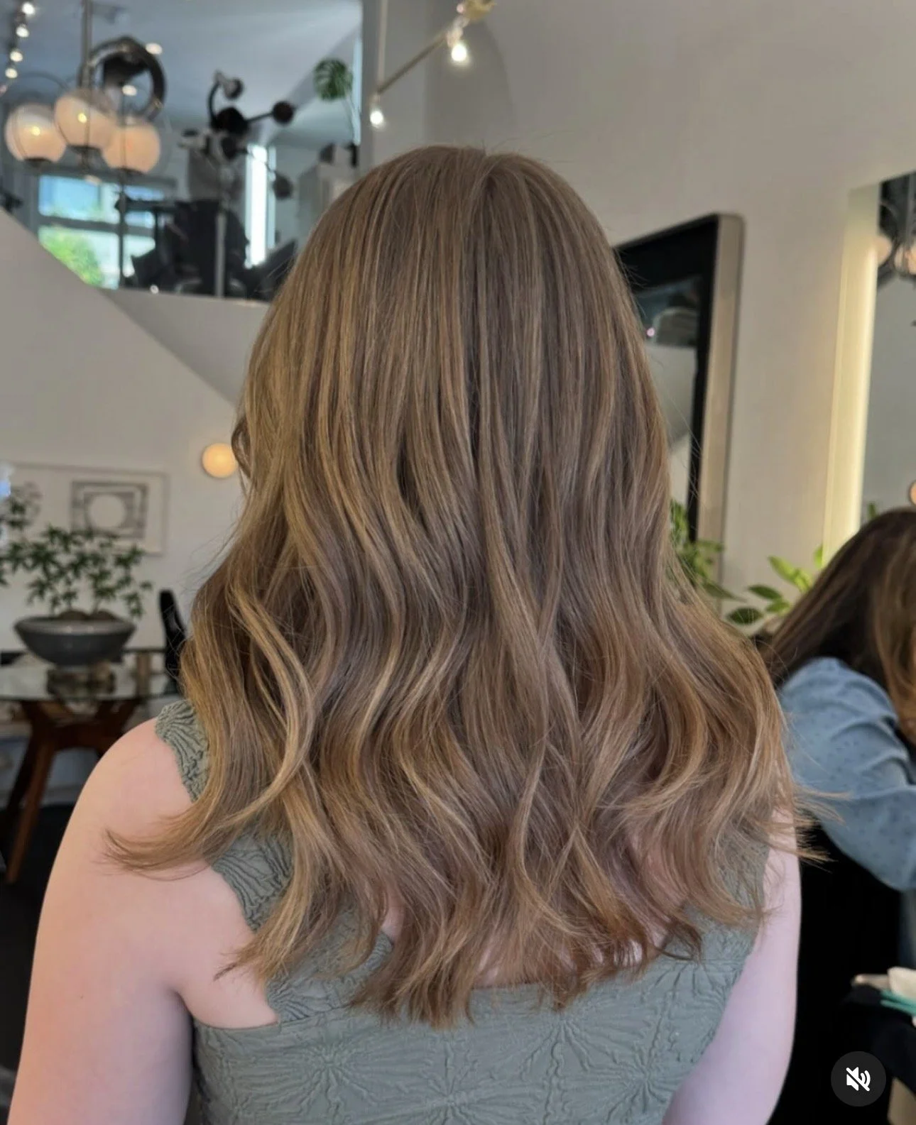 Back of a woman with long, wavy, light brown hair wearing a green sleeveless top in a modern indoor setting.