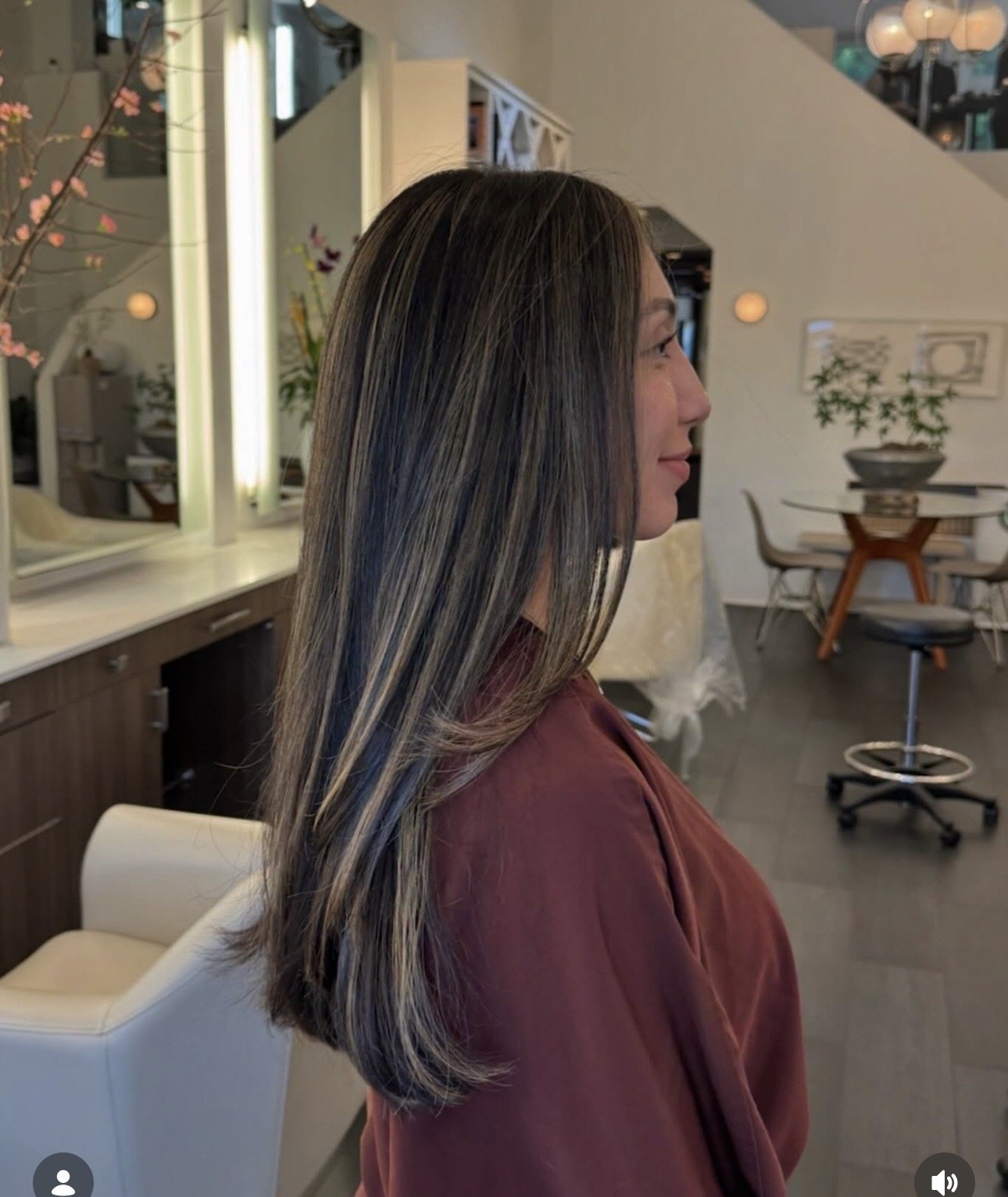 Side profile of a woman with long, straight, dark hair with blonde highlights, seated in a modern interior space.