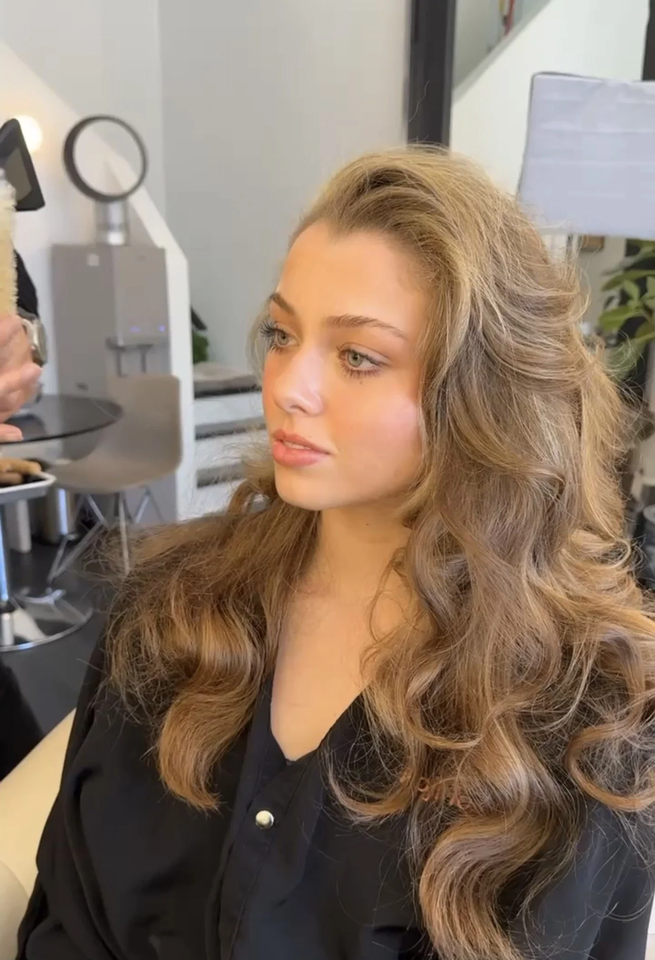 A woman with long, wavy, light brown hair and light skin getting her hair styled at a salon.