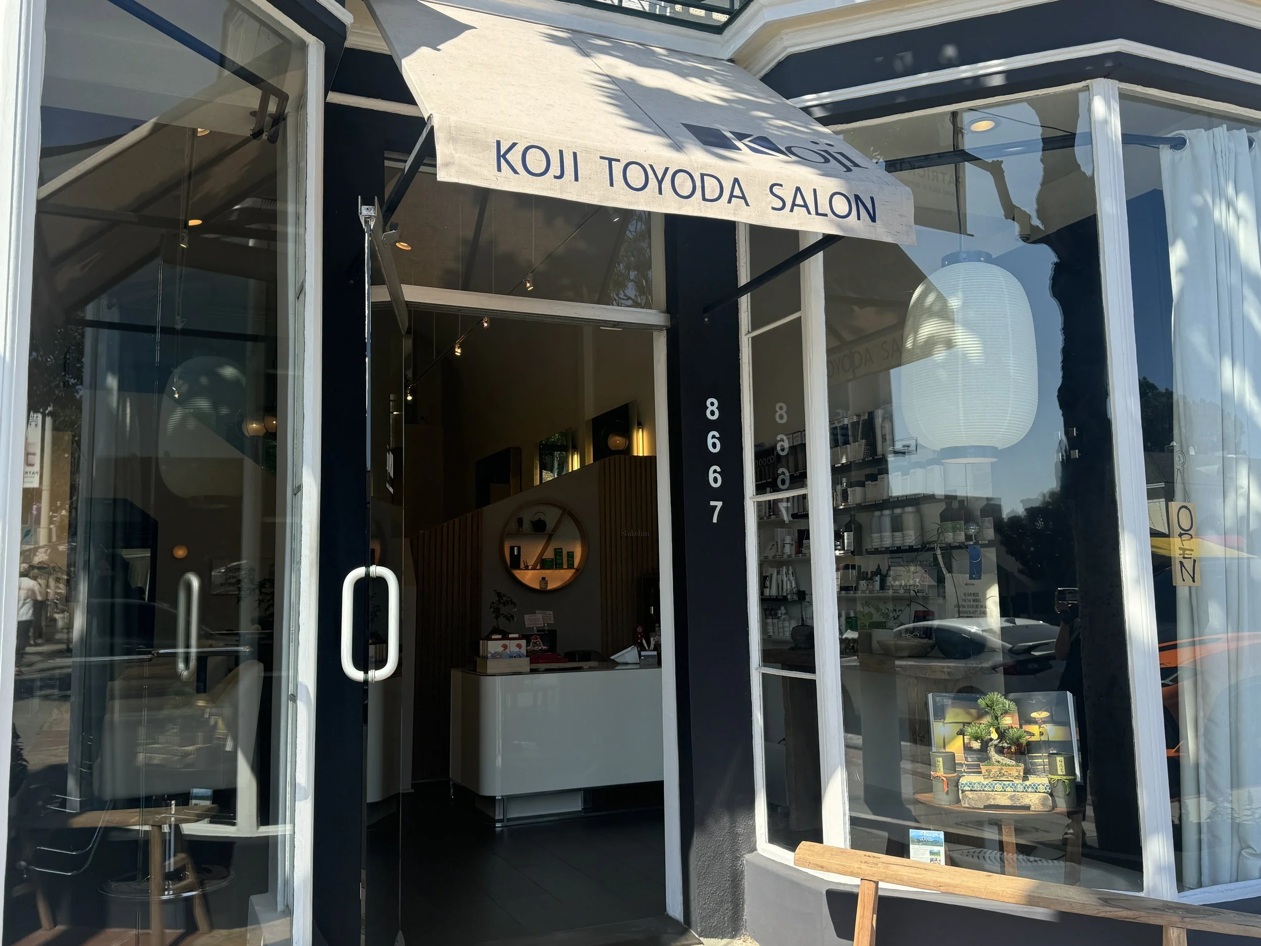 storefront of Koji Toyoda Salon with glass doors, navy blue exterior, and white signage, inside showing minimalistic interior with wooden accents and salon products, covered by an awning with the salon's name.