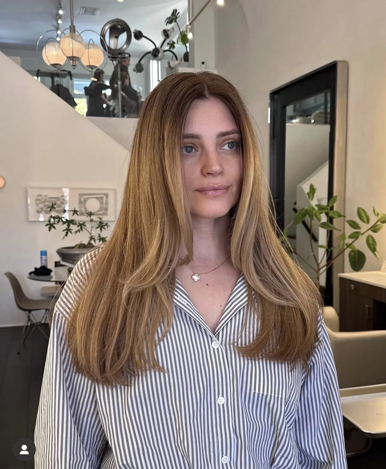 A young woman with long red hair and blue eyes wearing a striped shirt, standing inside a modern, well-lit cafe or restaurant with plants and contemporary decor.