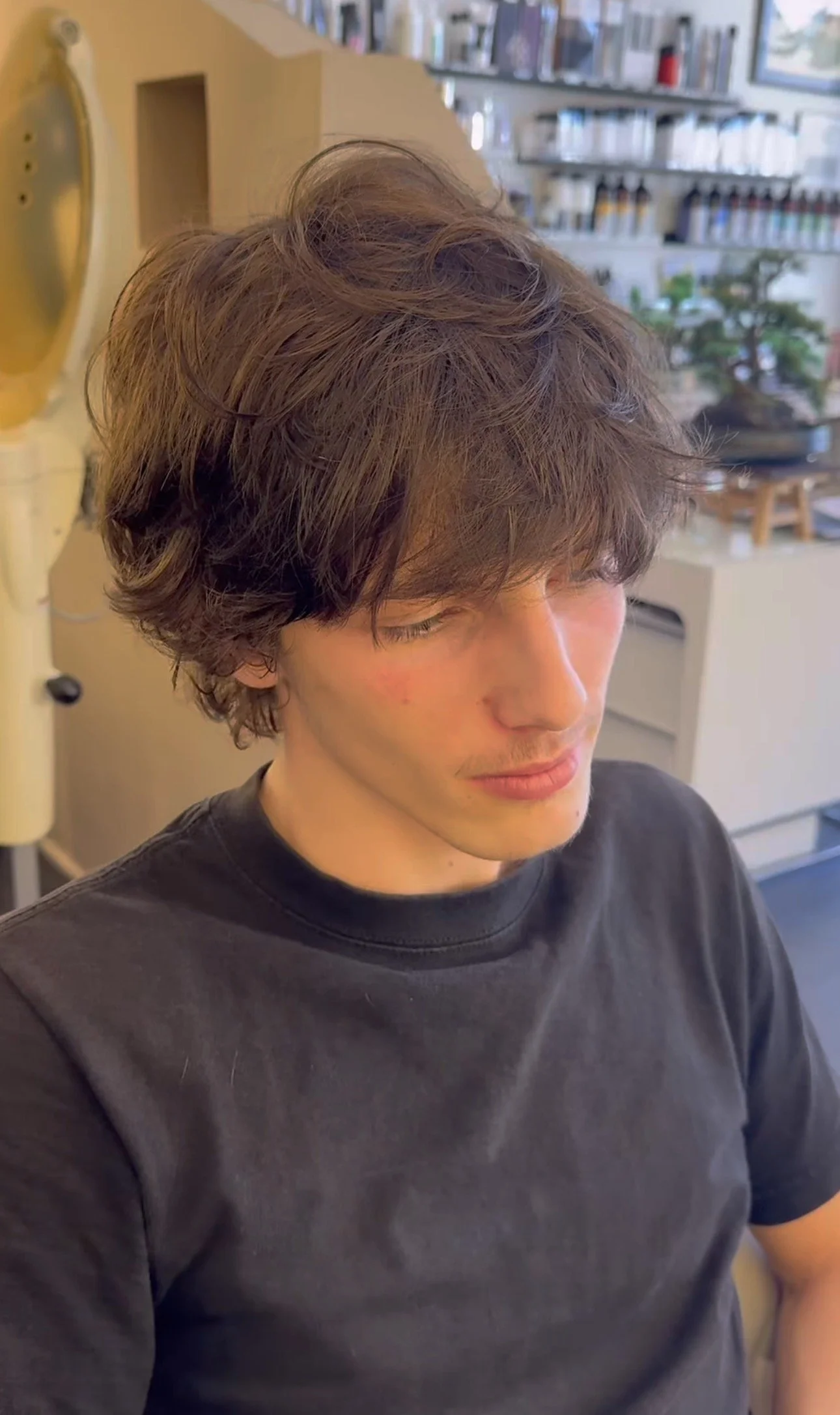 A young man with tousled brown hair and fair skin, wearing a black t-shirt, sitting indoors with bookshelves and decorative items in the background.