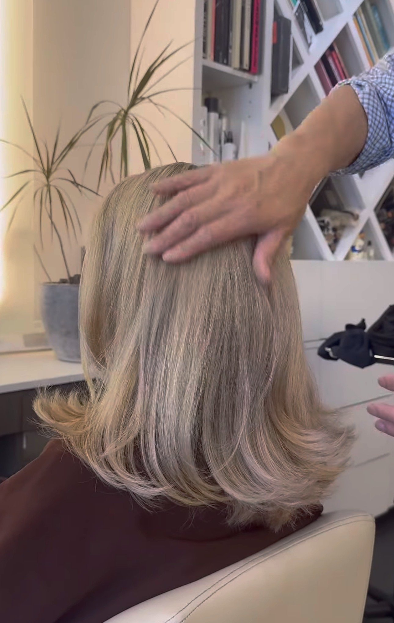 A person with blonde hair is getting a hair treatment at a salon, with a hand possibly applying a product or styling the hair, with a bookshelf and a plant in the background.