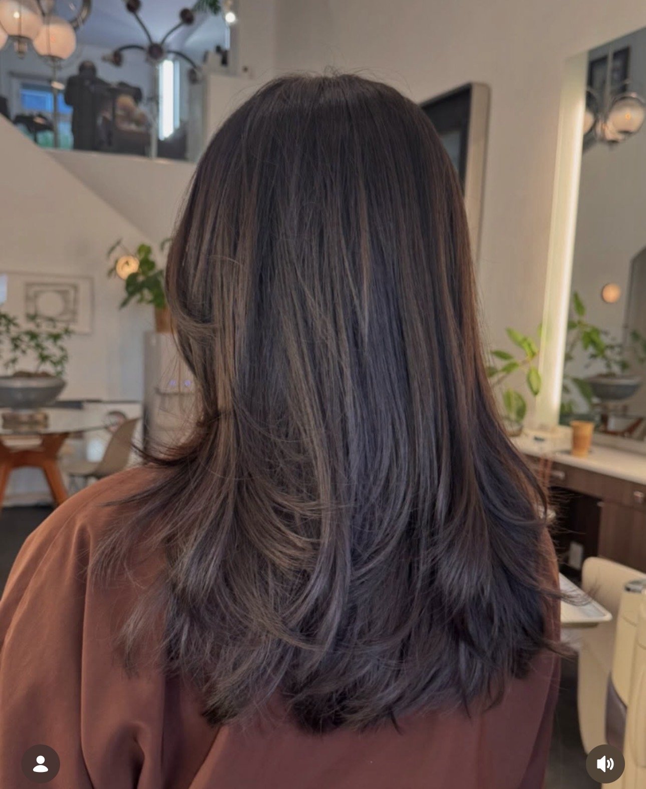 Back view of a woman with shoulder-length dark brown hair styled with loose waves, wearing a brown top in a cozy, well-decorated room.