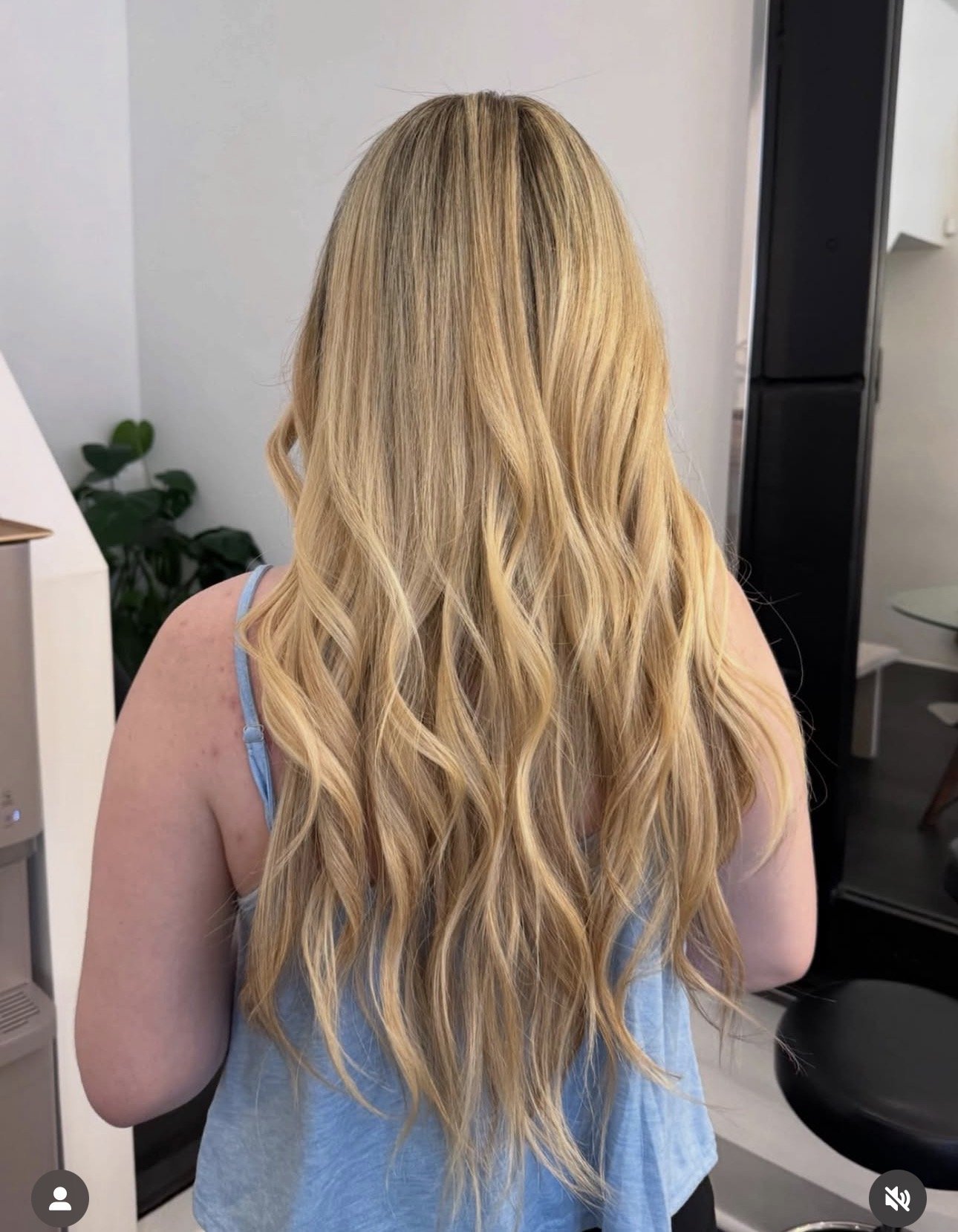 Back view of a woman with long, wavy blonde hair wearing a light blue top in a modern room with white walls and a black refrigerator.