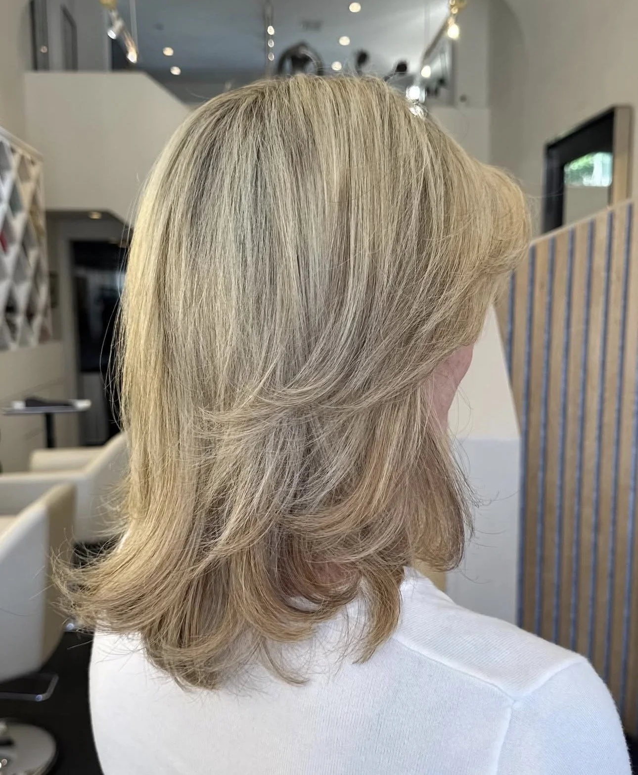 Back of a woman with shoulder-length, blonde hair styled in loose waves, wearing a white shirt, in a modern salon or office setting.