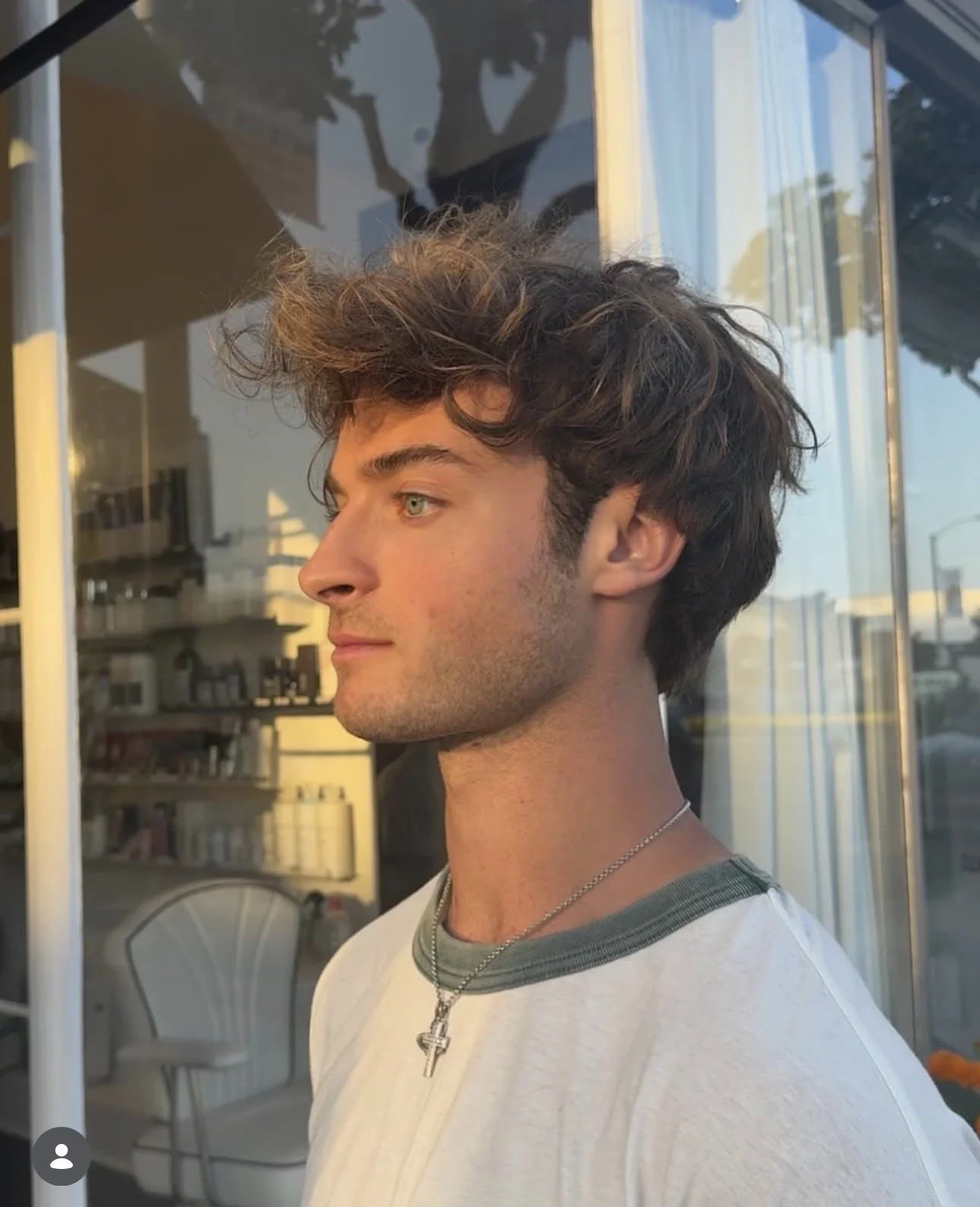 A young man with wavy brown hair and blue eyes looking out a window during sunset, wearing a white t-shirt with green trim and a silver cross necklace.