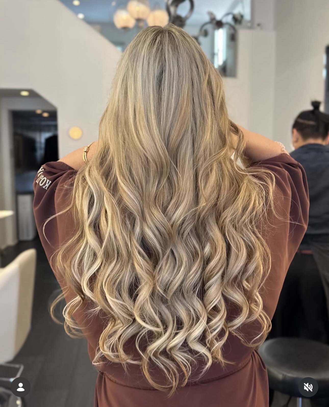Back view of a woman with long, blonde, wavy hair wearing a brown top, in a salon or similar setting.