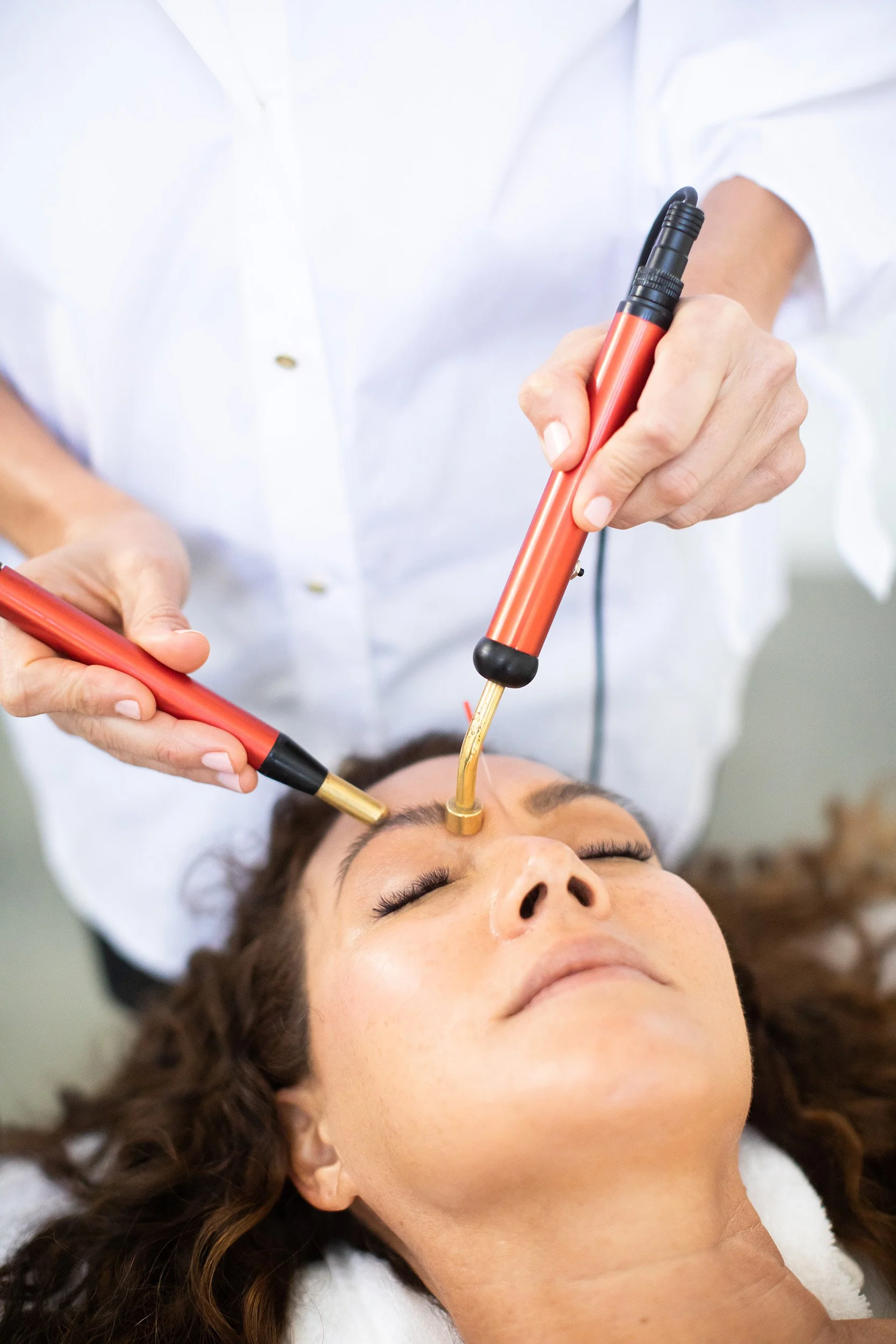A woman receiving a facial treatment with electrical devices held by a practitioner.