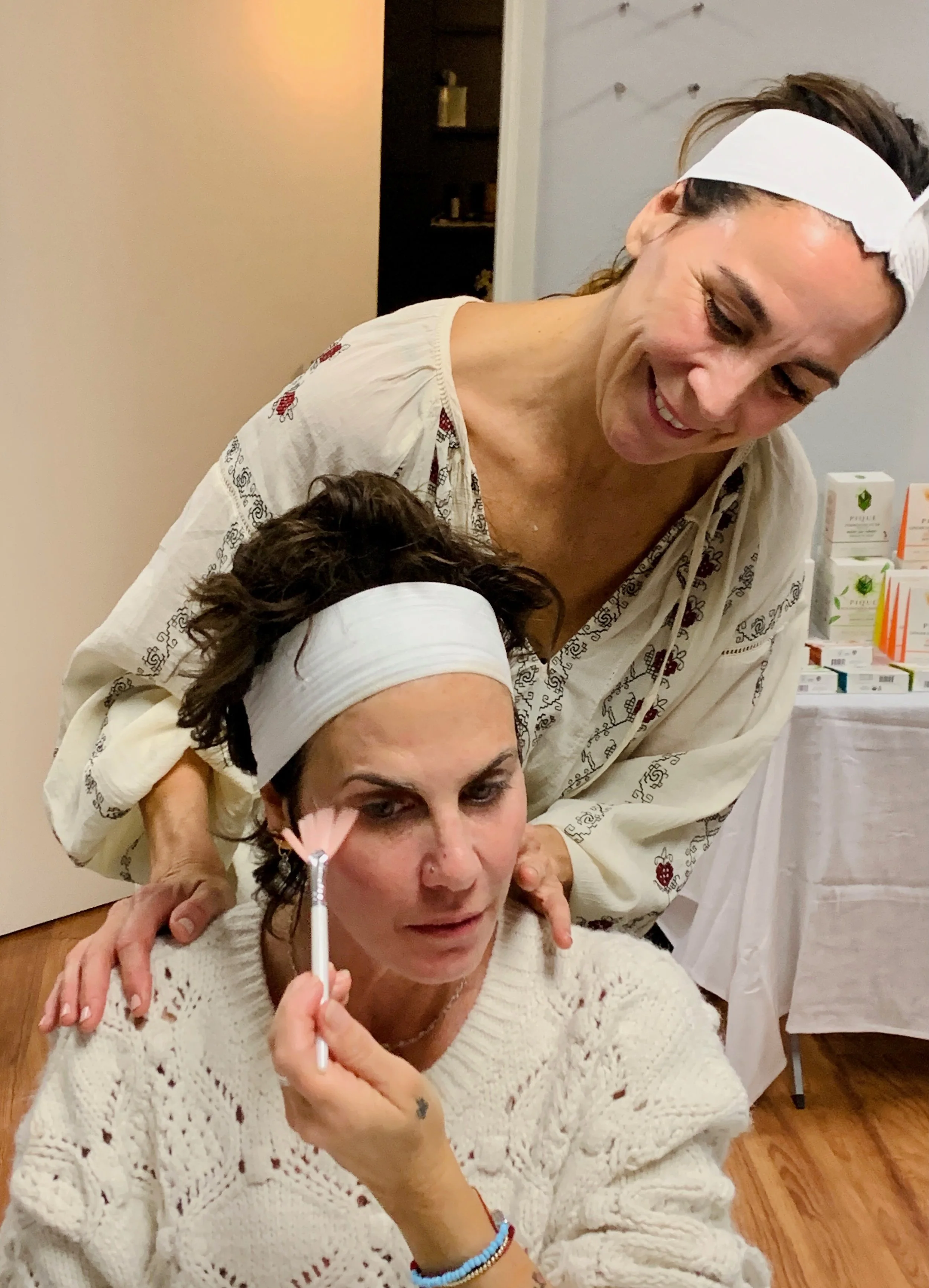 Two women, one seated and one standing behind her, are engaged in a skincare or cosmetic session. The seated woman is wearing a white headband and a chunky knit sweater, applying a facial mask or treatment with a small brush. The standing woman is smiling, wearing a white headband and a patterned blouse, assisting with the skincare process. A table with skincare products and boxes is visible in the background.