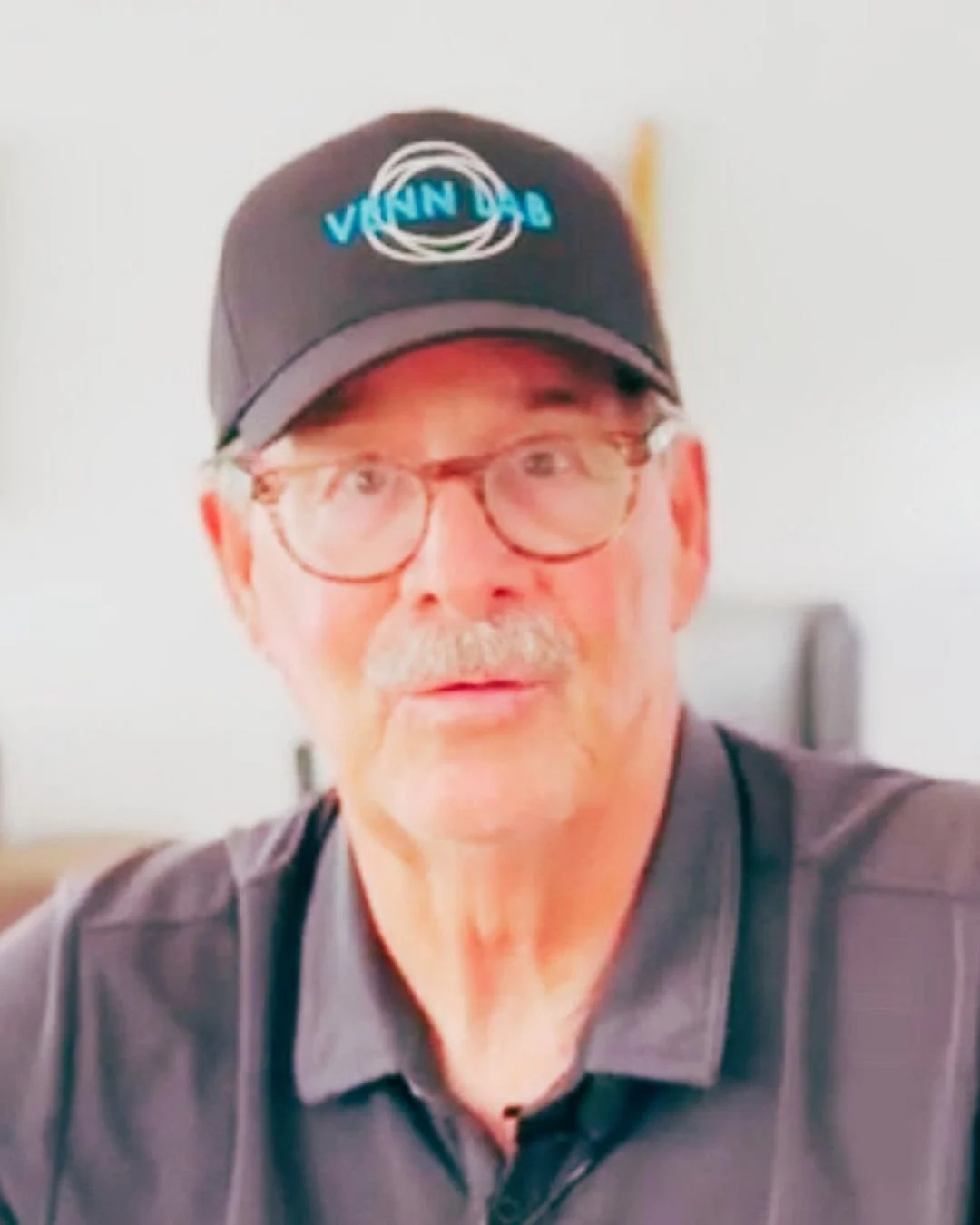 A man with glasses, a mustache, and a beard wearing a black cap with blue and white logo, and a dark gray collared shirt.