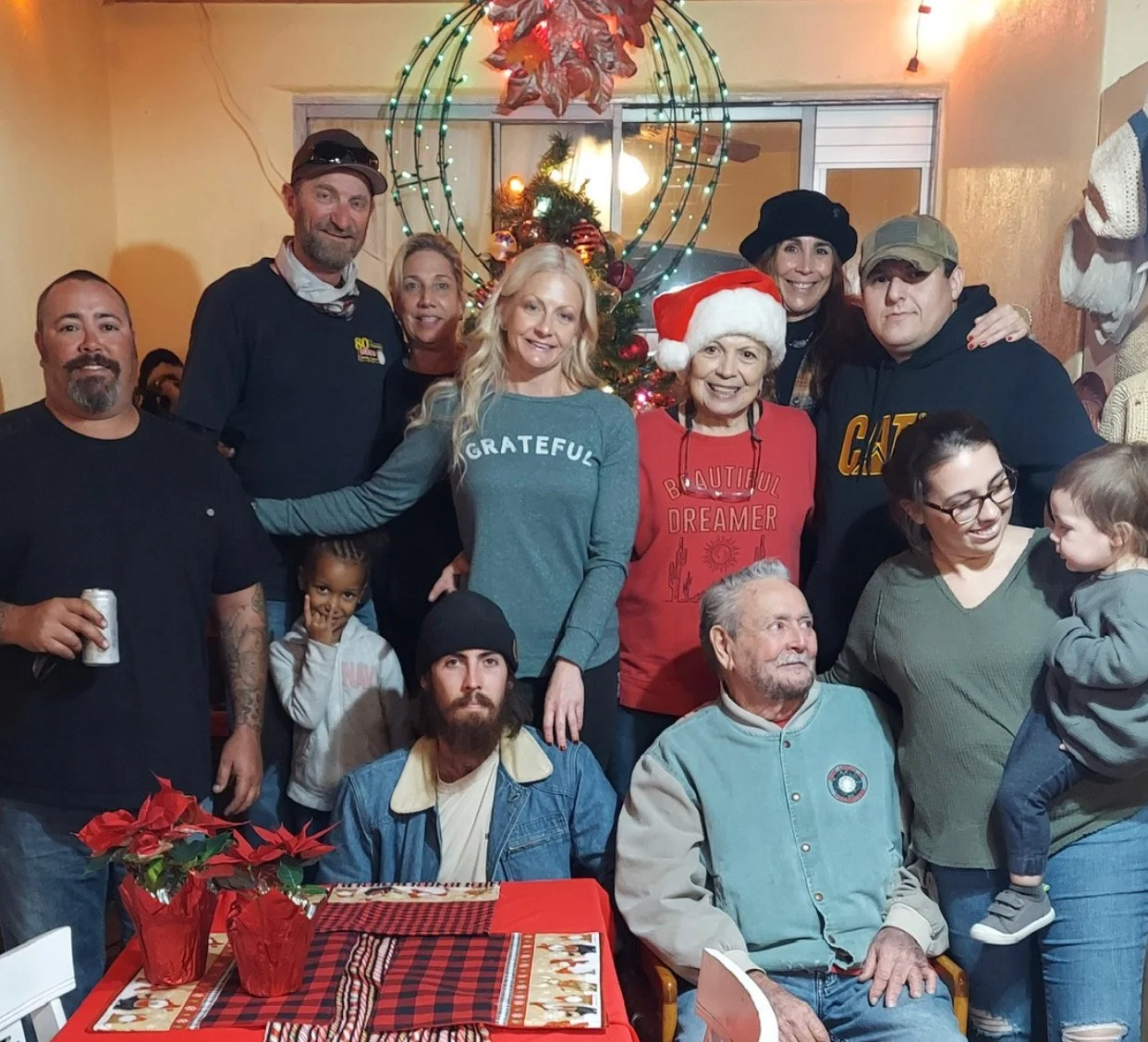A group of people gather around a Christmas tree, smiling, with some wearing Christmas-themed clothing, including a woman with a Santa hat and a red shirt. The scene is festive, with holiday decorations and a table with poinsettias and a tablecloth in the foreground.
