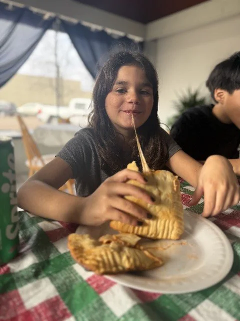 A girl smiling while pulling melted cheese from a grilled cheese sandwich at a dining table.