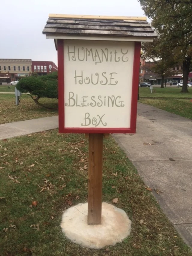 A sign on a wooden post in a grassy area that reads 'HUMANite HOUSE BLESSING BOX'. The sign has a shingle roof and is placed near a sidewalk with trees and buildings in the background.