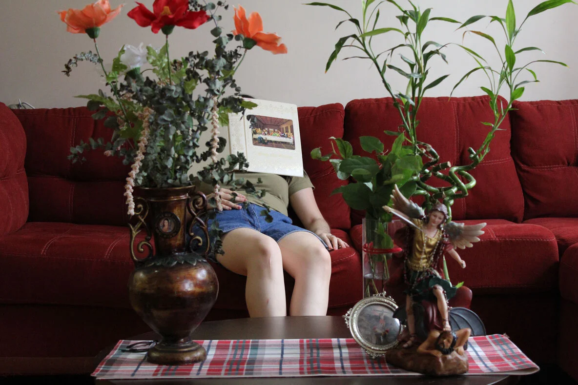 Delaney Diaz-Tapia's photos in the Studio Museum Harlem Hearts in Isolation exhibition An art photo of a red couch with a young person leaning back on it with a book over their face. In the foreground is a large, tall bouqet and an angel statue on the coffee table.