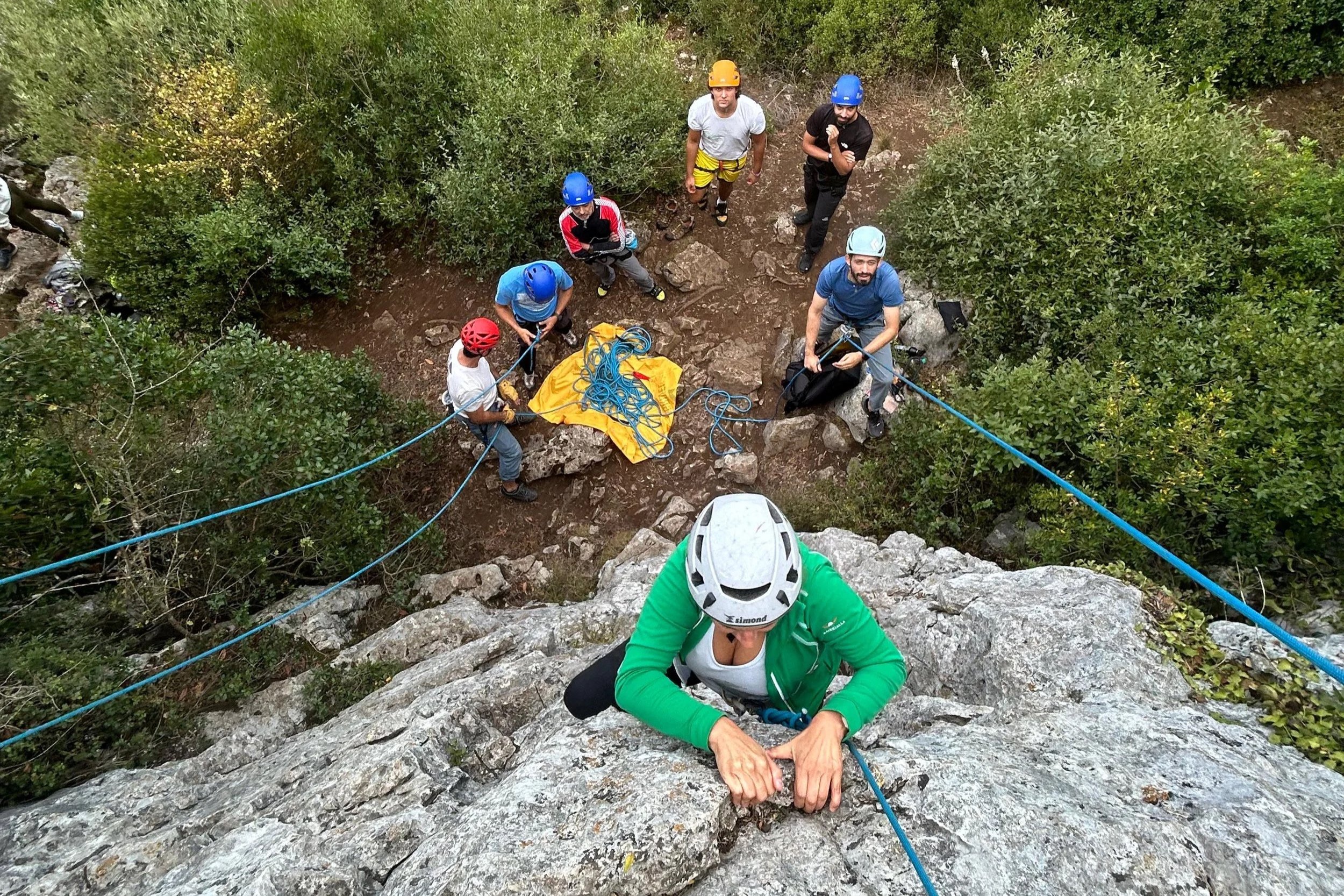 Pessoa escalando uma rocha com grupo de pessoas ao redor, usando capacete, com vegetação ao redor.