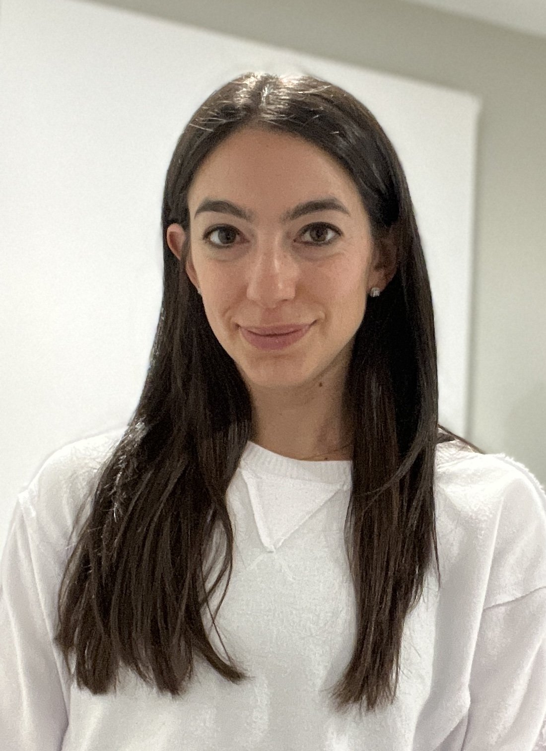 A woman with long dark brown hair smiling, wearing a white sweater, standing indoors against a plain background.