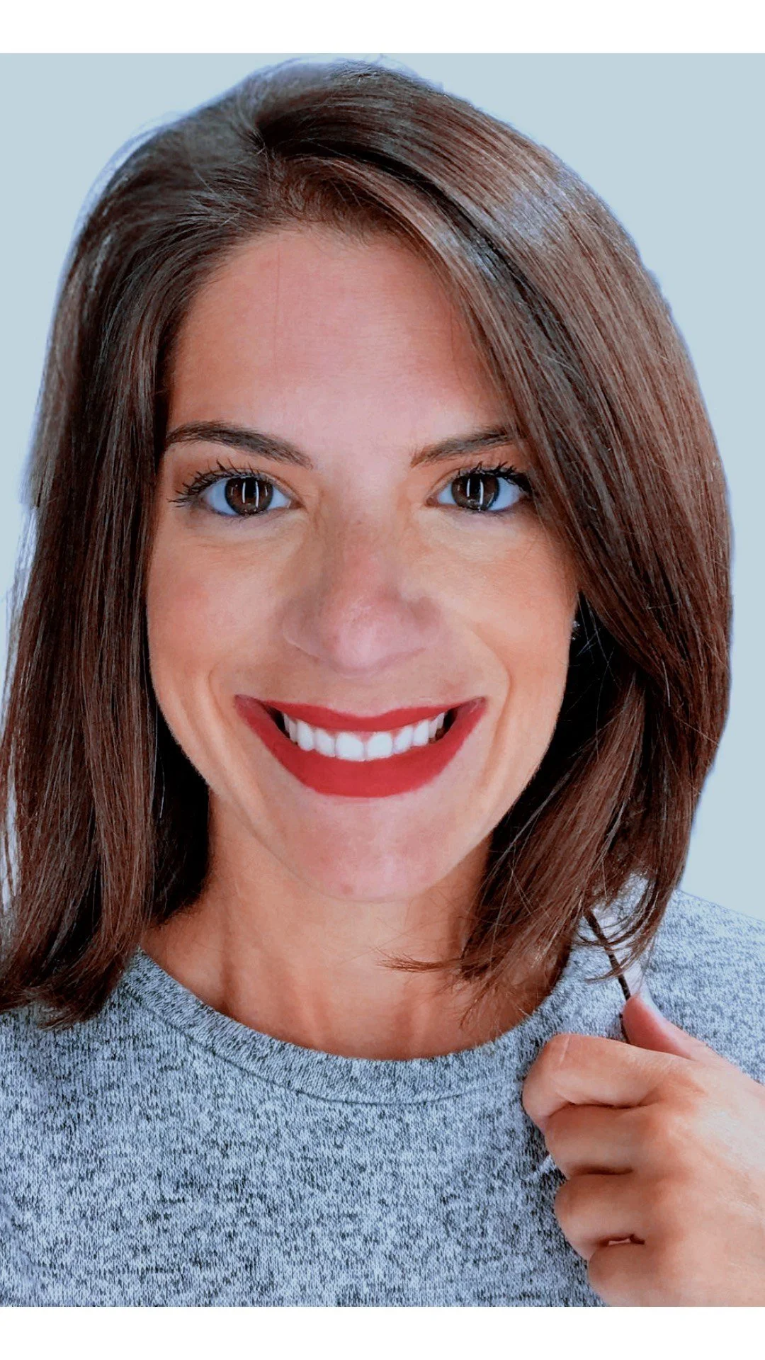 Close-up of a smiling woman with shoulder-length brown hair, blue eyes, and wearing a gray shirt, holding a strand of her hair.