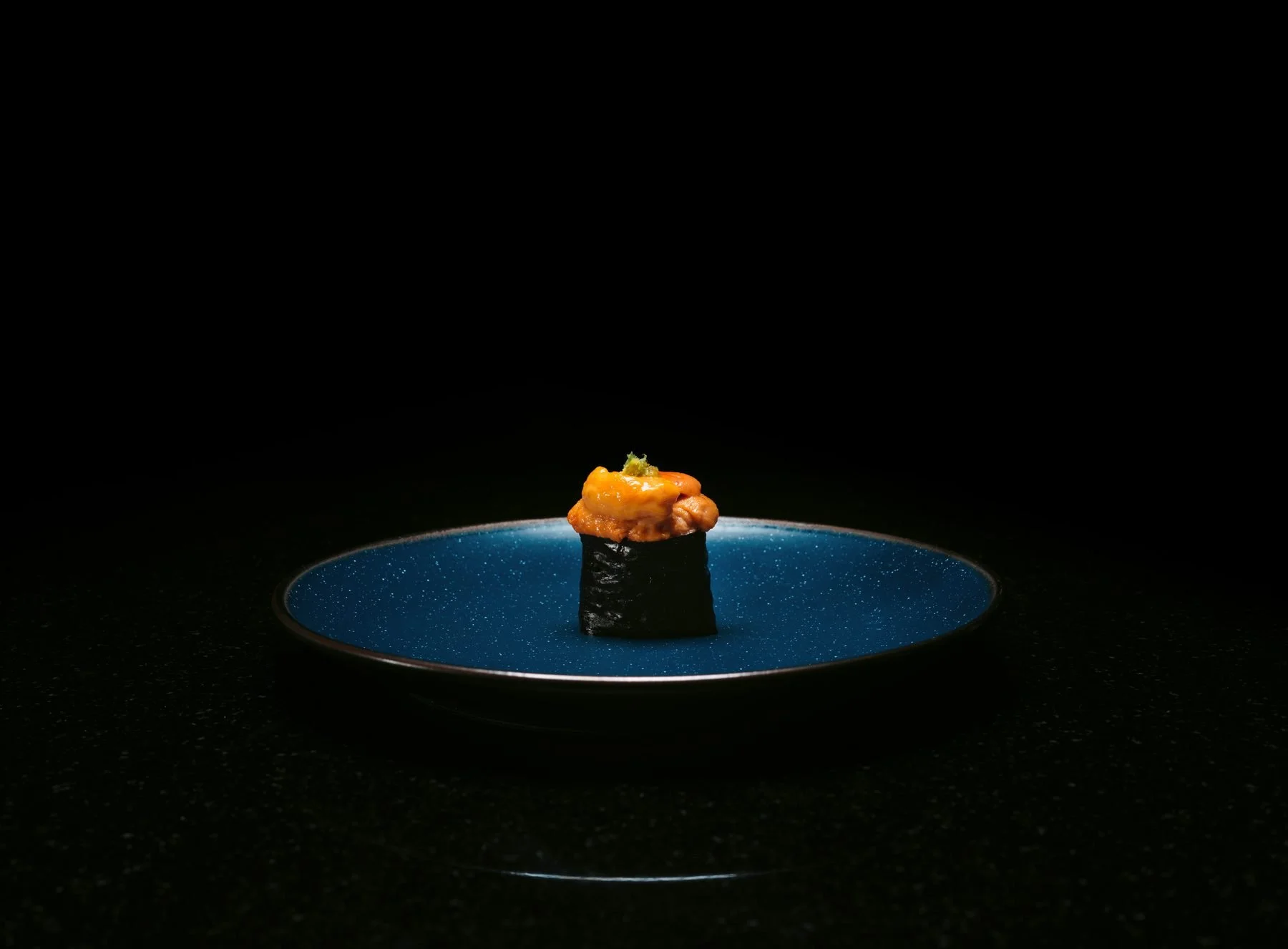 A piece of sushi topped with orange fish roe and green garnish, placed on a dark plate with a blue speckled interior, against a black background.