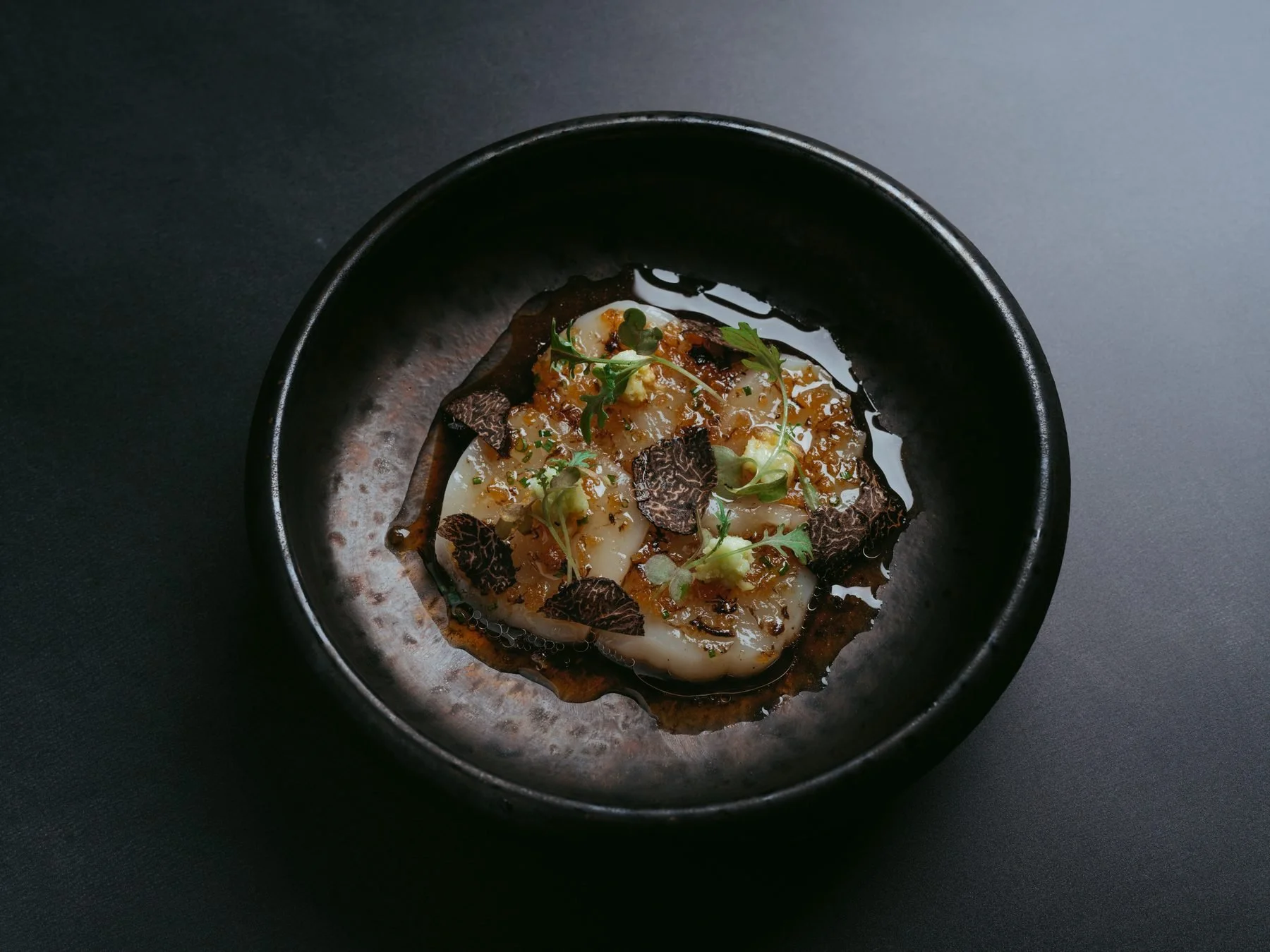 Dish of ravioli pasta in a dark bowl, topped with microgreens, black truffle slices, and a sauce garnished with herbs.