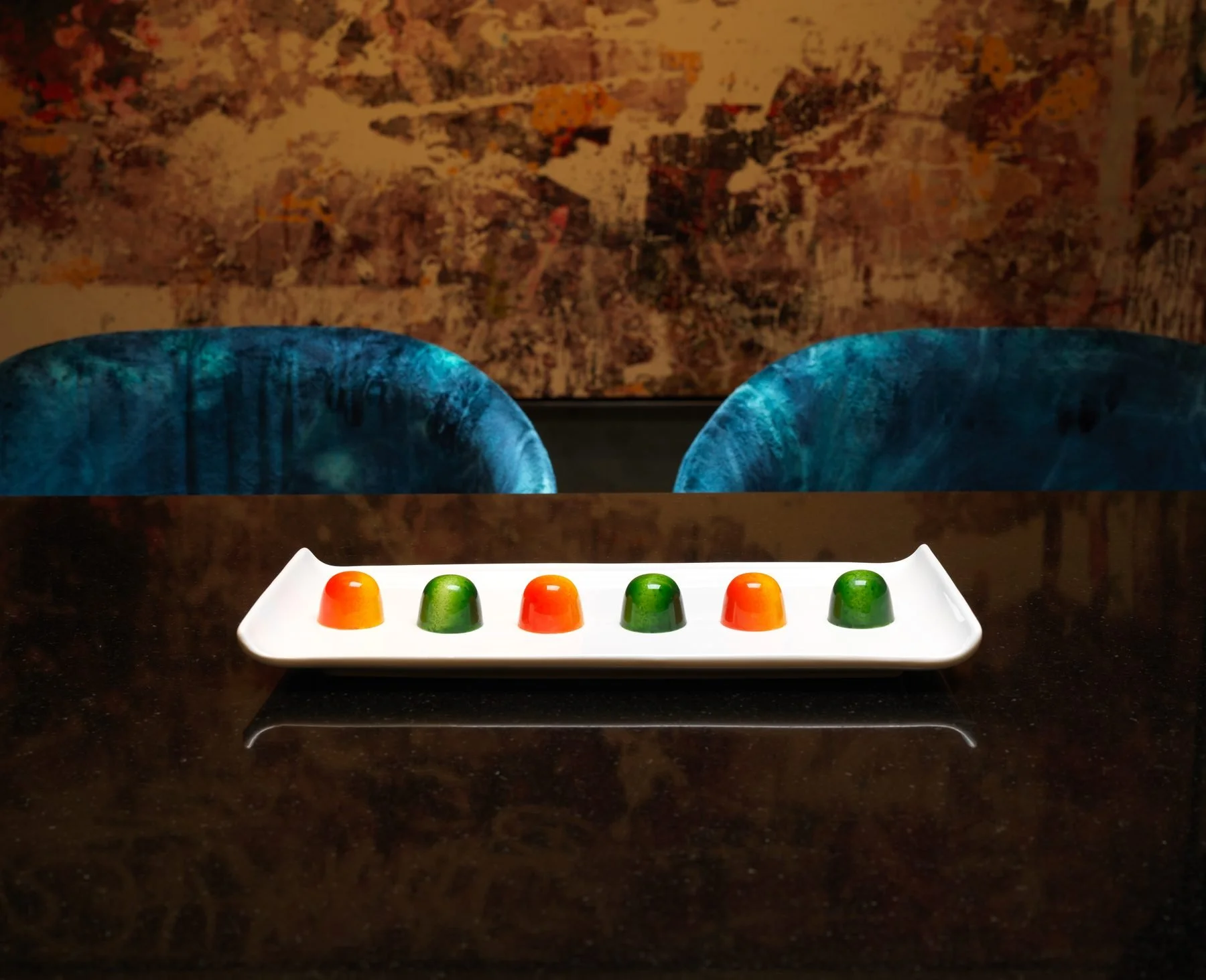 A white rectangular tray with six colored jelly candies arranged in a row, featuring orange, green, orange, green, orange, and green candies, placed on a dark countertop. In the background, there are two blue velvet chairs with curved backs and a textured abstract wall art with shades of brown, orange, and purple.
