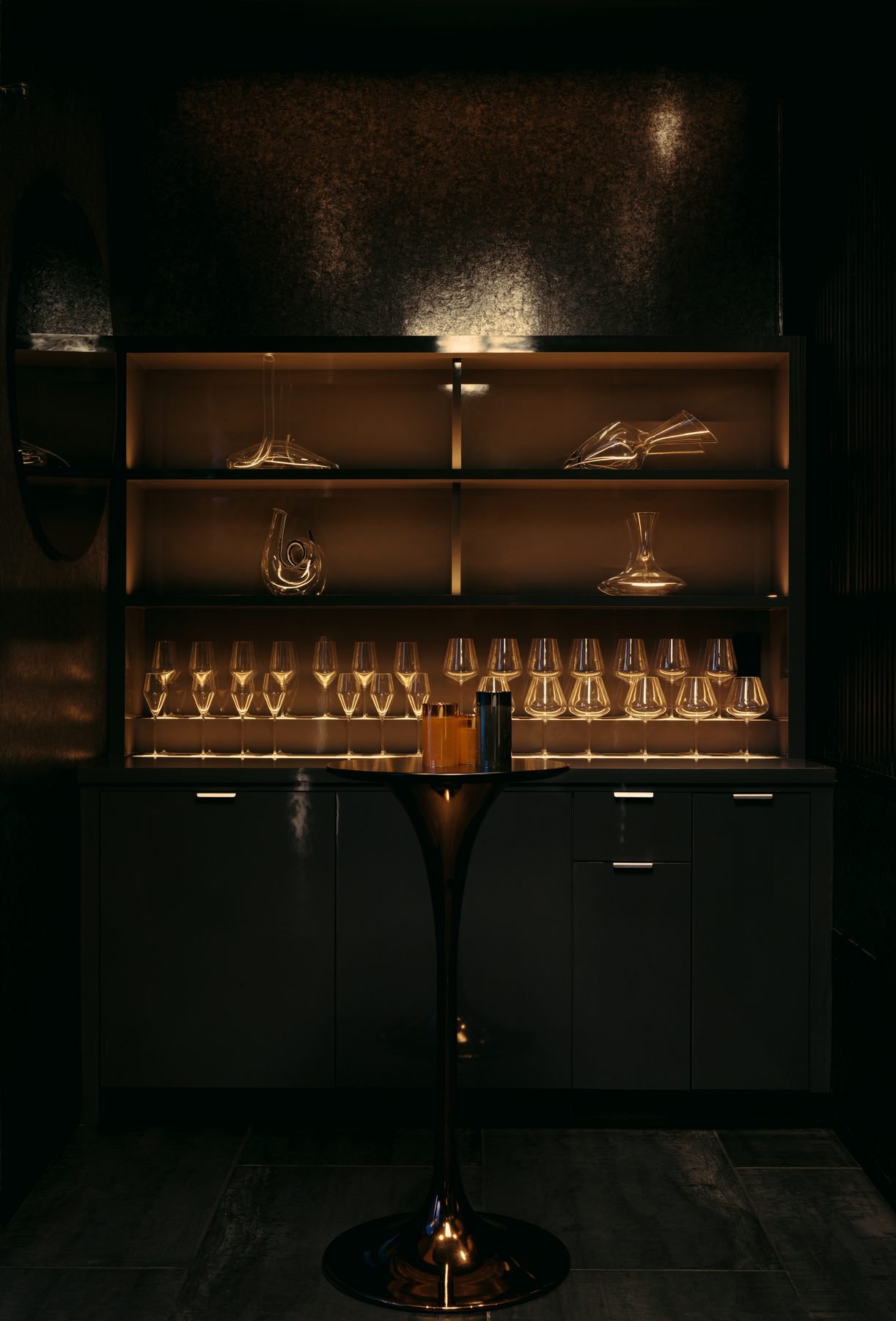 A black cabinet with glassware on shelves, illuminated by warm lighting, with a black table and metallic vases in front, set against a dark textured wall.