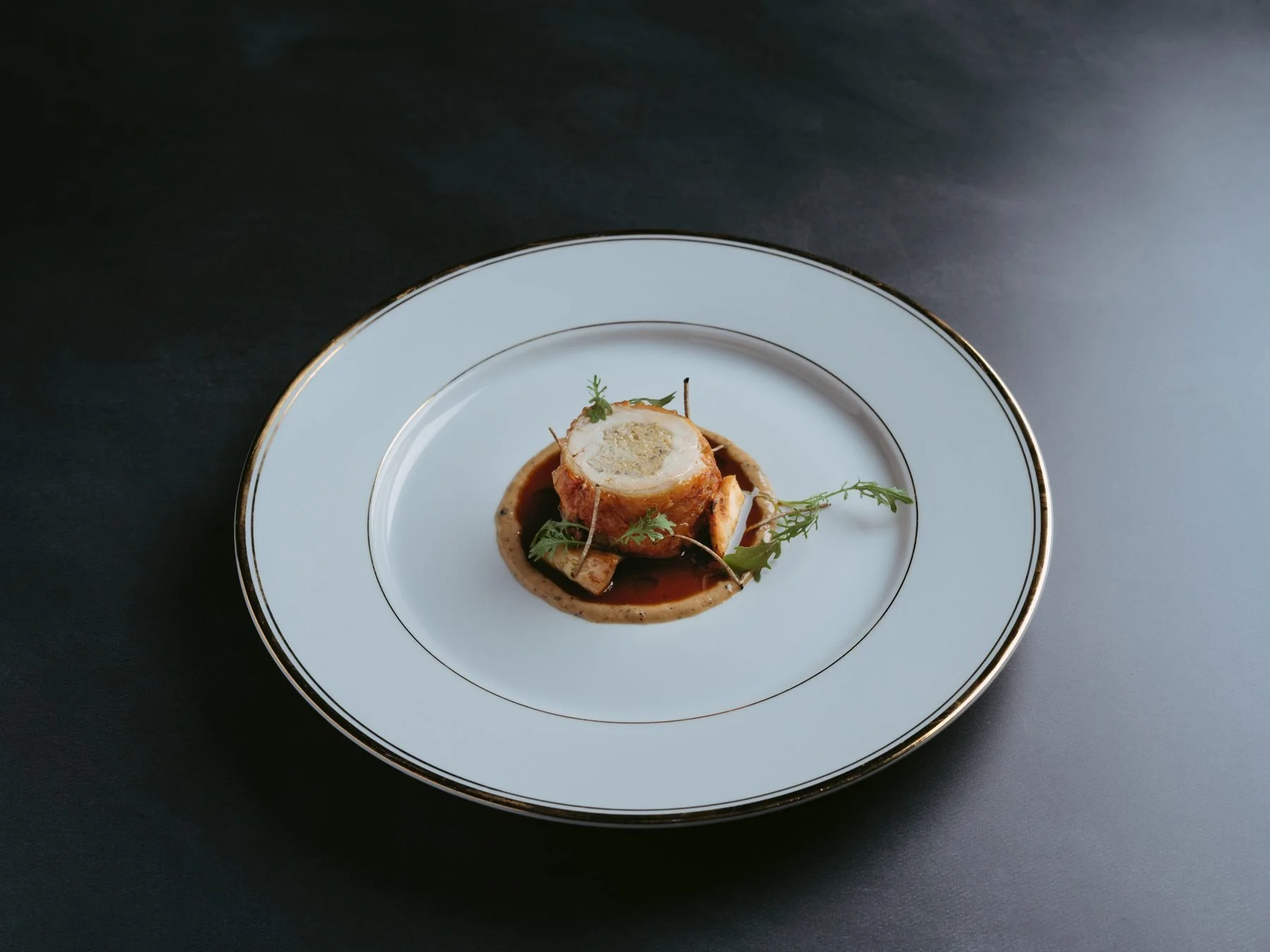 A plated gourmet dish with a wrapped roulade, garnished with herbs, served on a white plate with gold trim, on a dark surface.