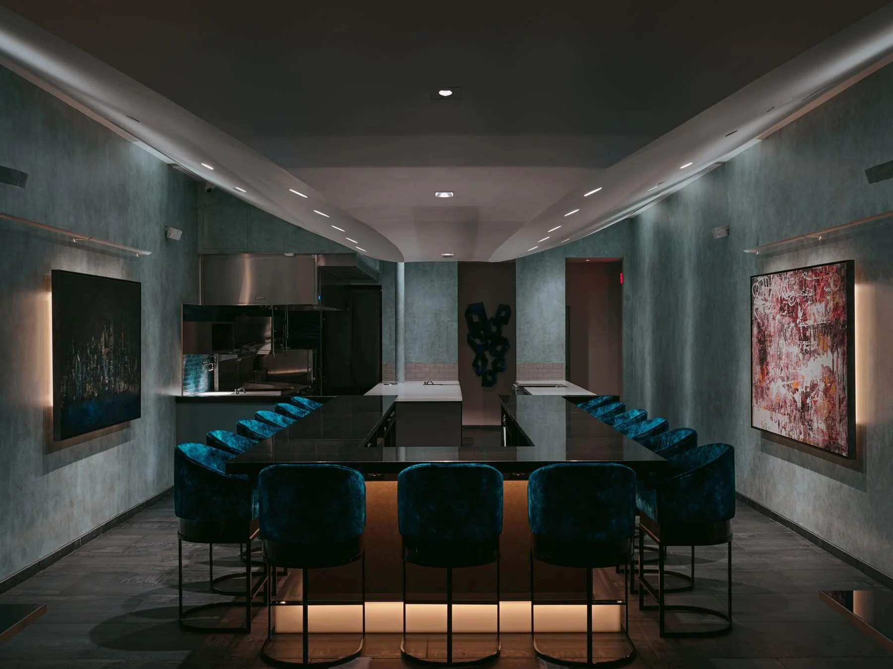 A modern dining area with a long black table surrounded by blue velvet chairs. The walls are decorated with abstract artwork, and there is a kitchen area visible in the background. The ceiling has installed lighting, and the overall ambiance is sleek and contemporary.