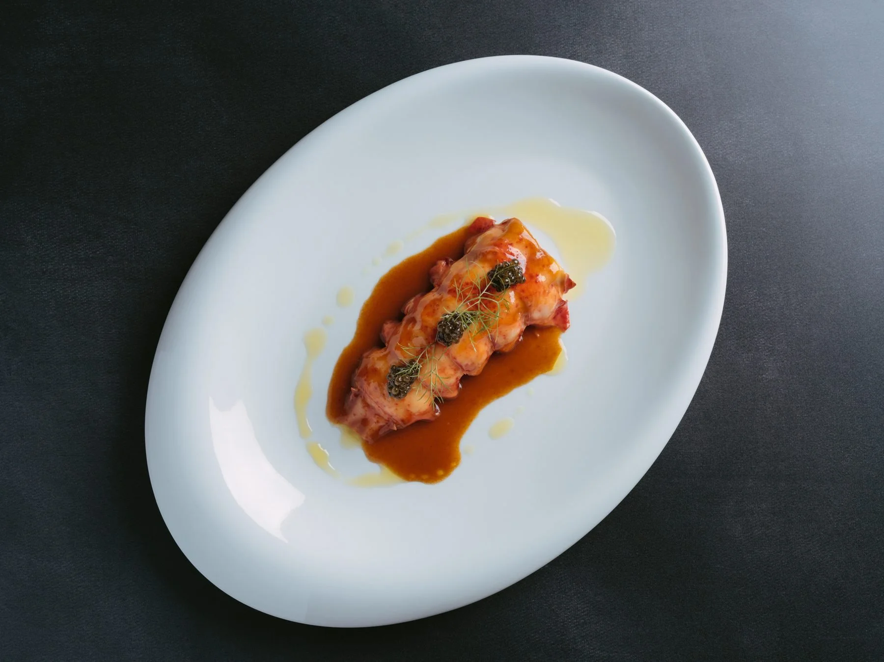 A plated dish featuring a rolled meat entrée topped with caviar and garnished with herbs, served with a brown sauce and a drizzle of oil on a white oval plate.