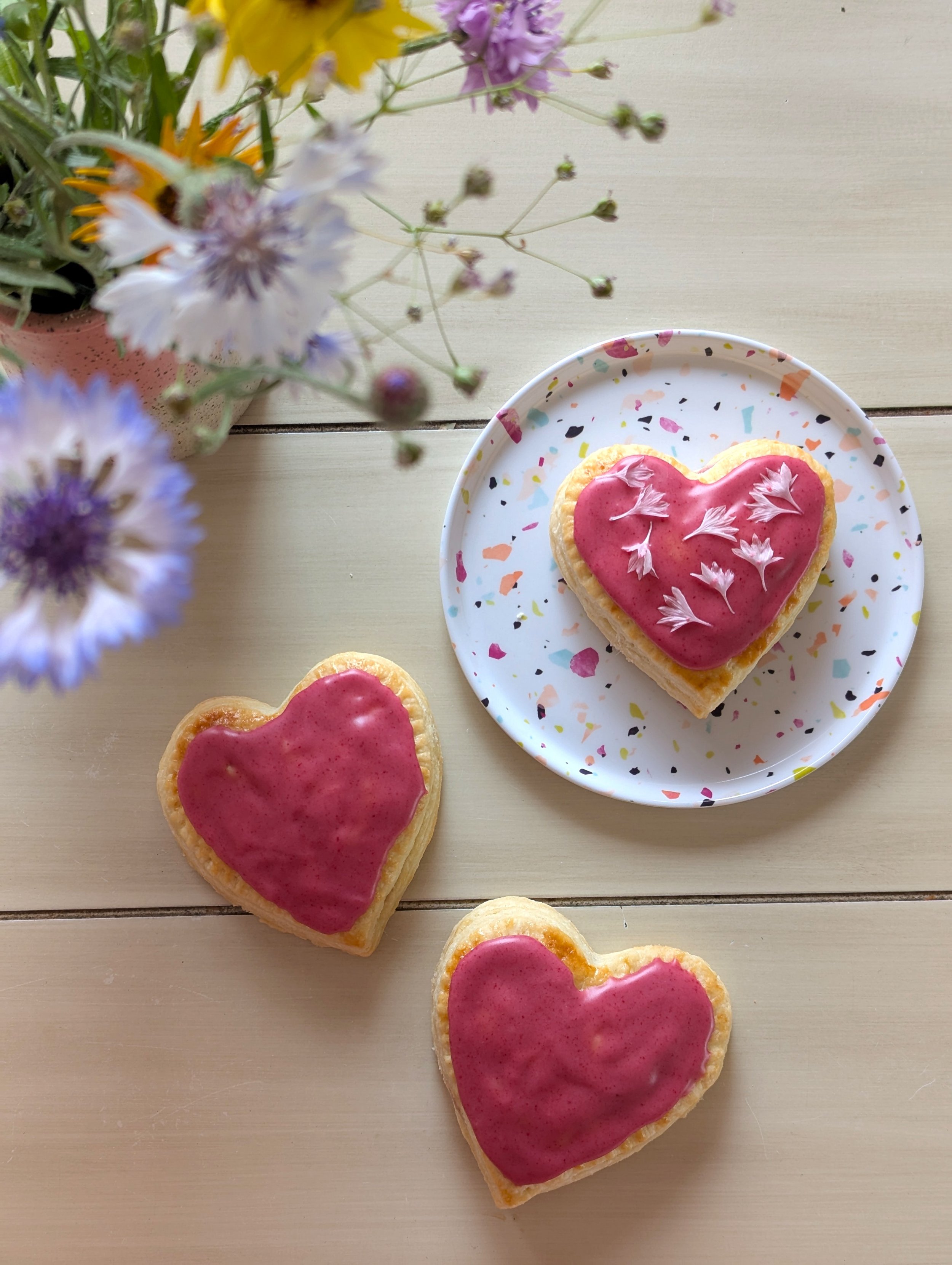 3 heart shaped raspberry hand pies with raspberry glaze sit on a tabletop beside a vase of colourful flowers