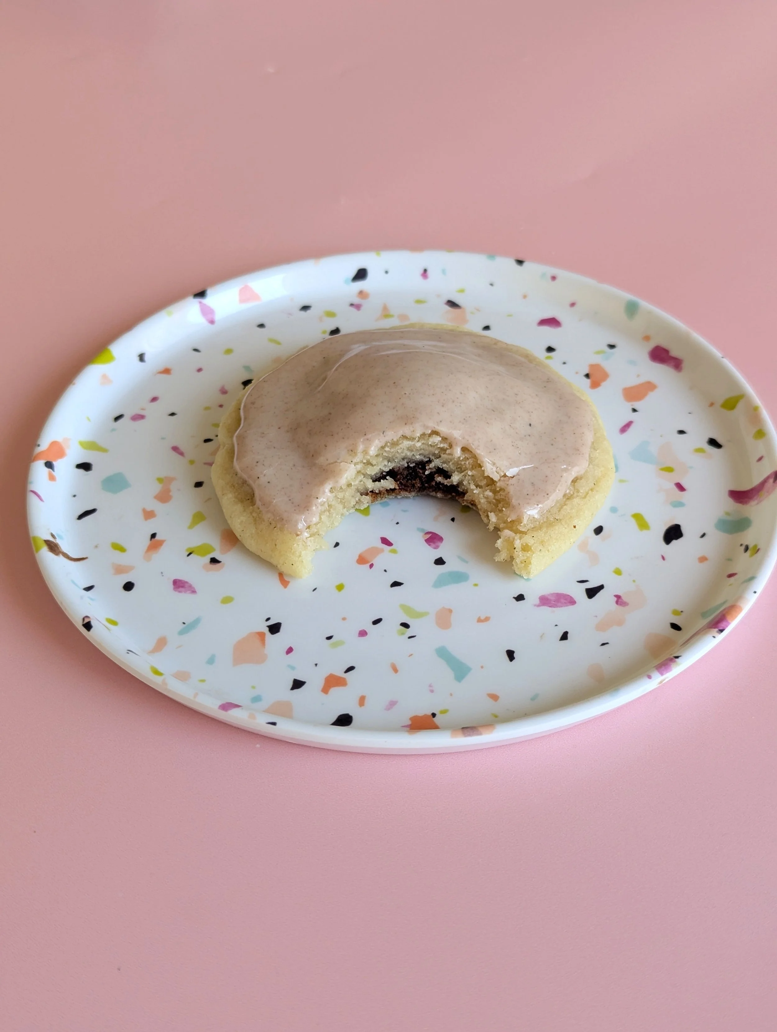 A glazed brown sugar pop tart cookie with a bit taken out of it sites on a small colourful plate on a pink background