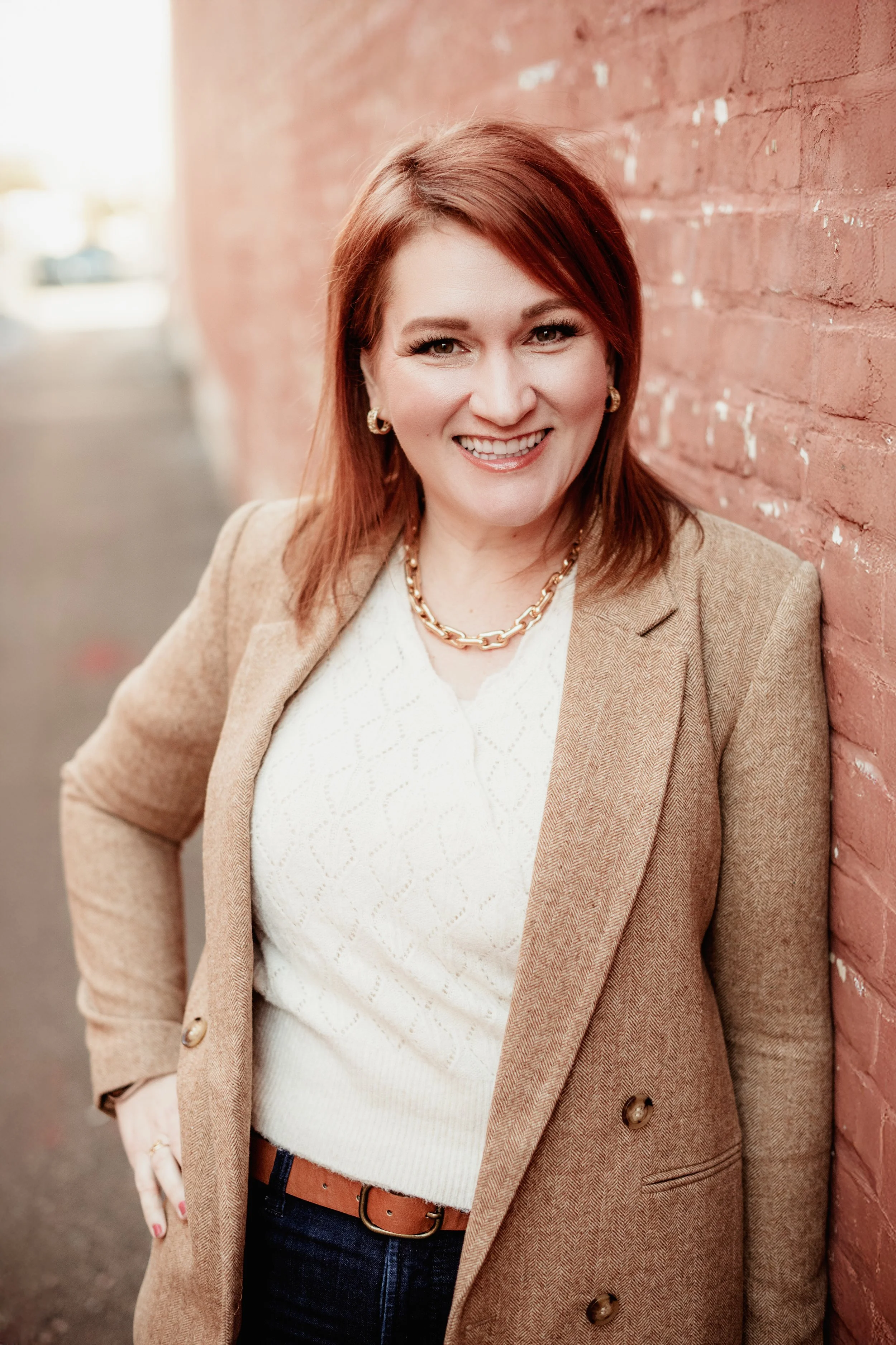 Woman with red hair in a suit jacket, leaning up against a red brick wall, smiling