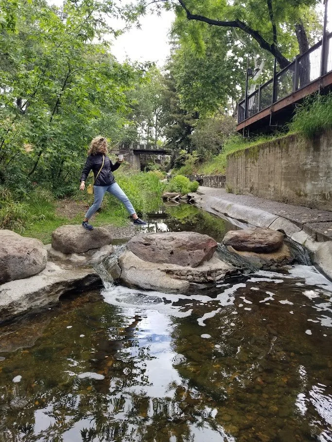Active woman in her 50s practicing mindful proprioception and functional balance by stepping across stones in a creek, illustrating Somatic Yoga Therapy for fall prevention in San Marcos.