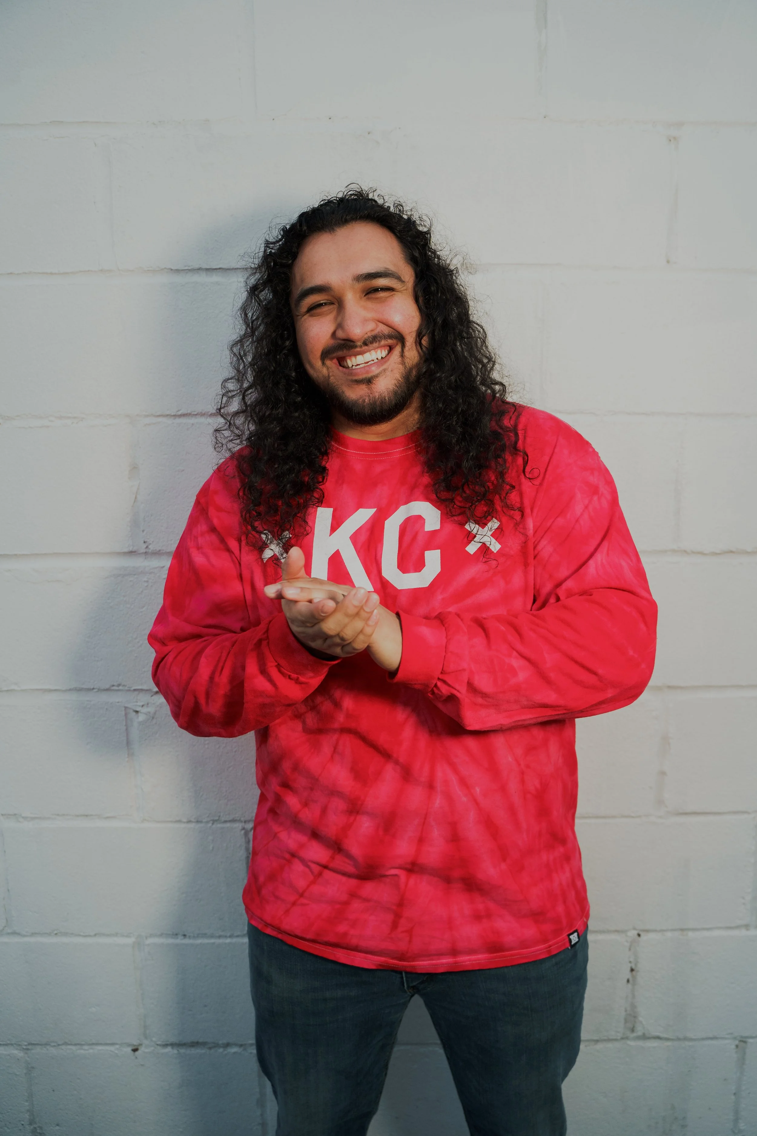 A man with long curly black hair and a beard smiling, wearing a red tie-dye sweatshirt with white letters, standing against a white brick wall.