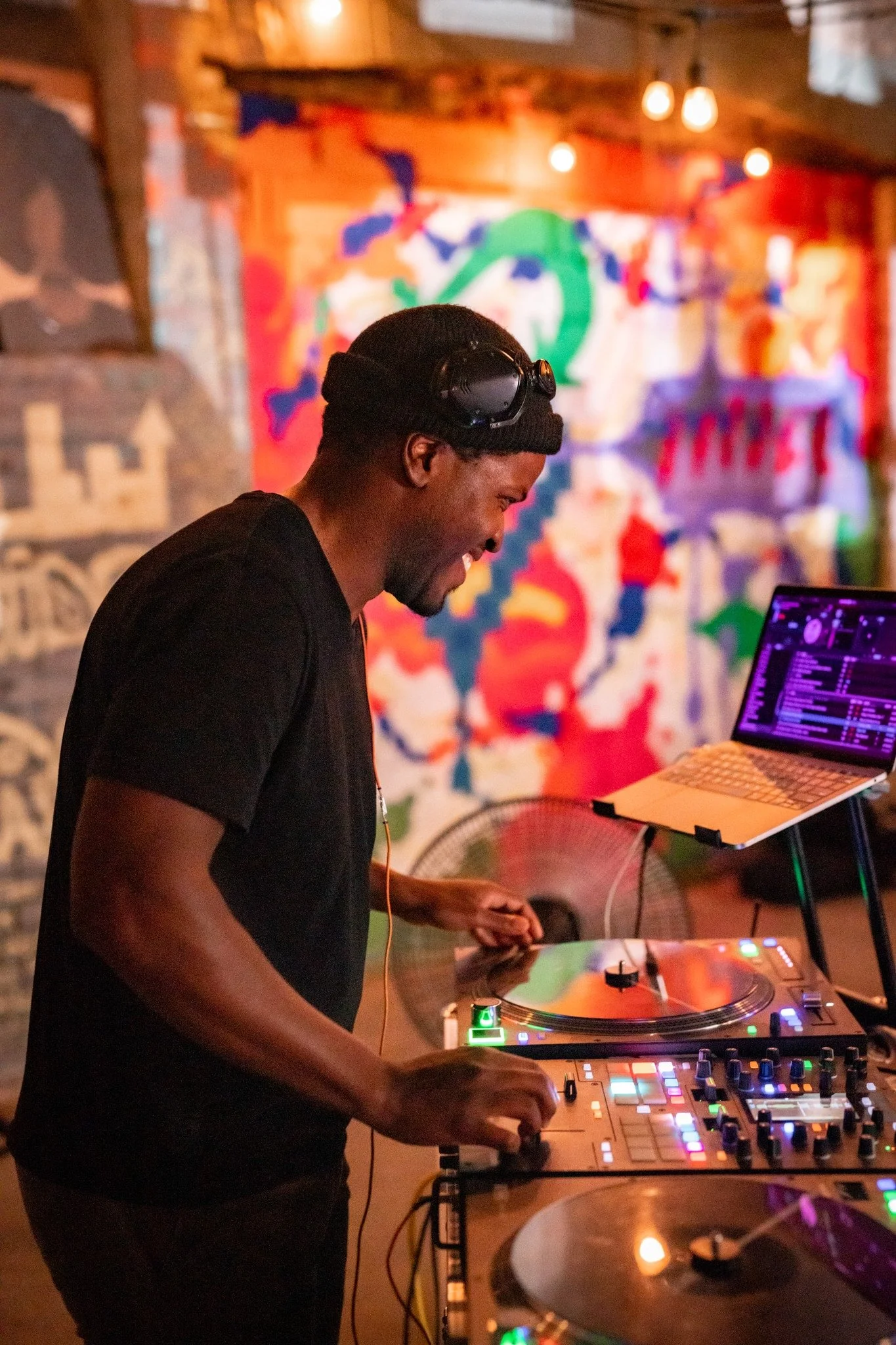 A person DJing with turntables and a laptop, in a dimly lit room with colorful graffiti on the wall in the background.